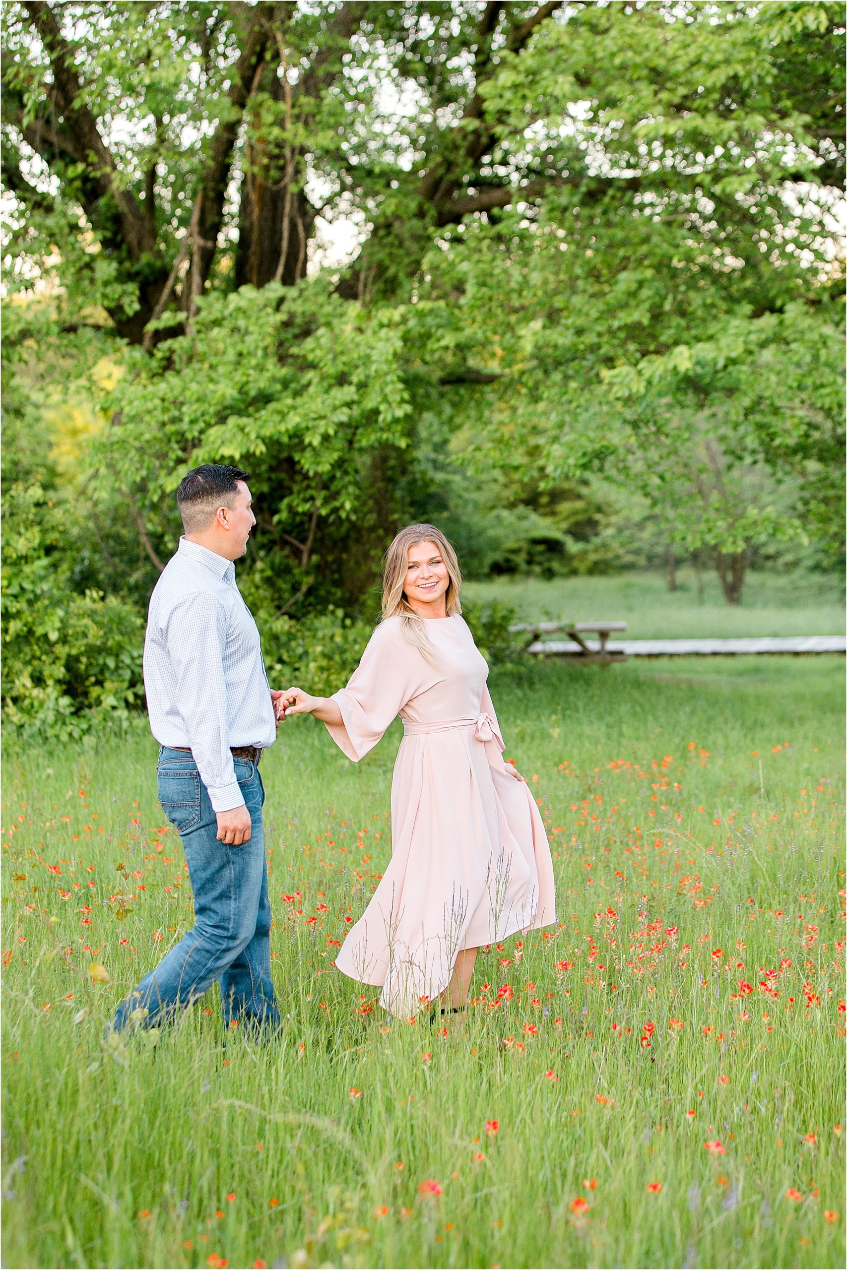 A bride to be holding hands with her fiance and leading him through a lush field for their engagement session in Fort Worth, Texas