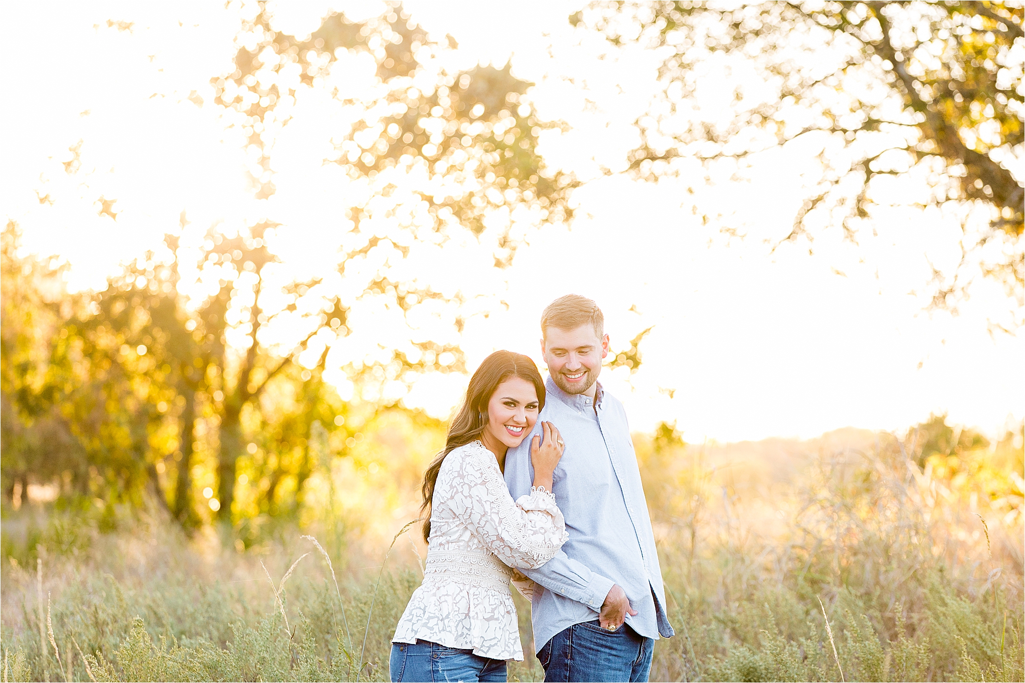 A groom peeks back over his shoulder while his fiance leans on his shoulder and smiles at the camera during their golden hour engagement session at a field in McKinney, Texas 