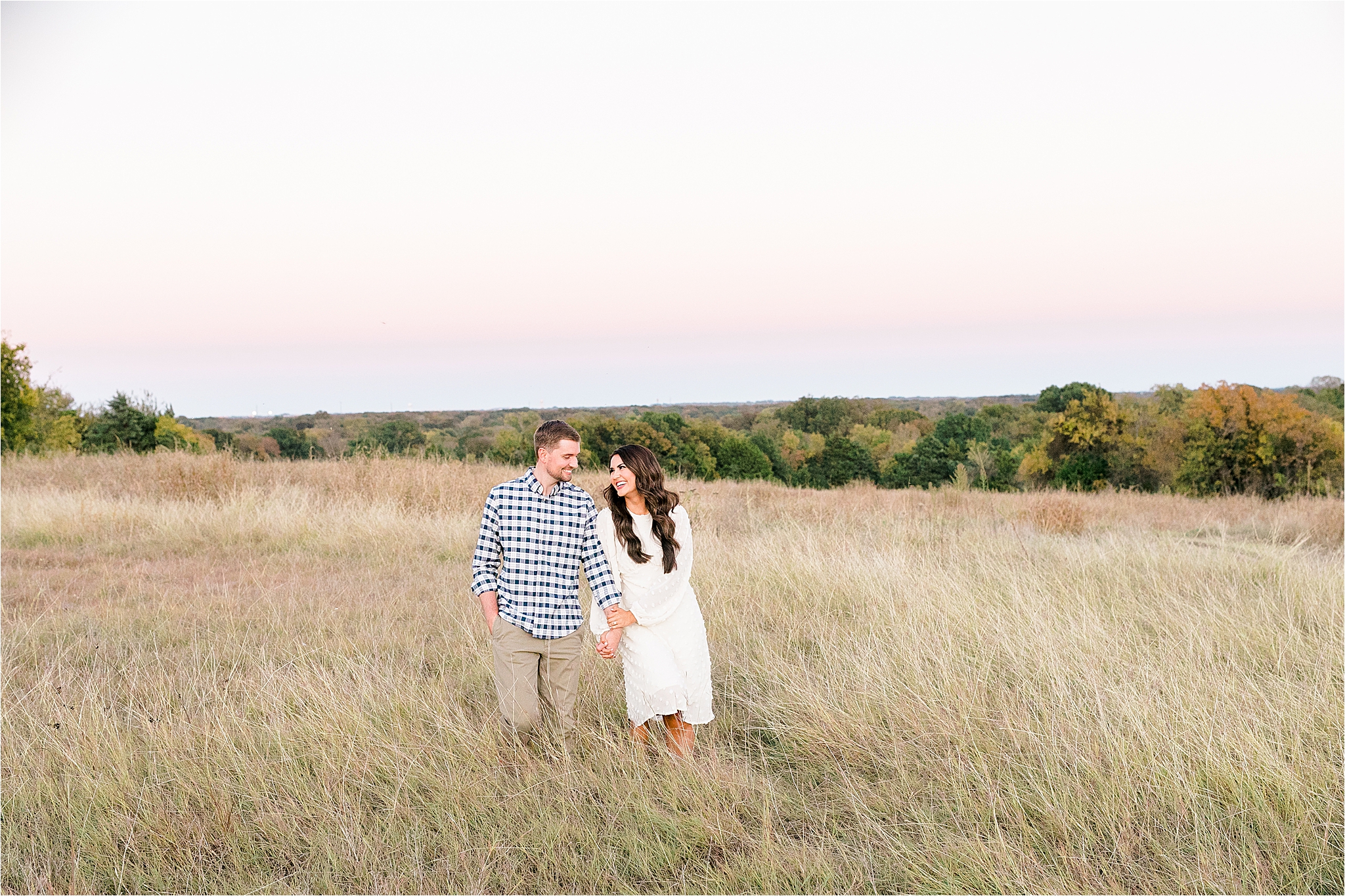 A brunette bride holds hands with her groom in a blue plaid shirt as they run though a field at sunset during their engagement session in McKinney, Texas