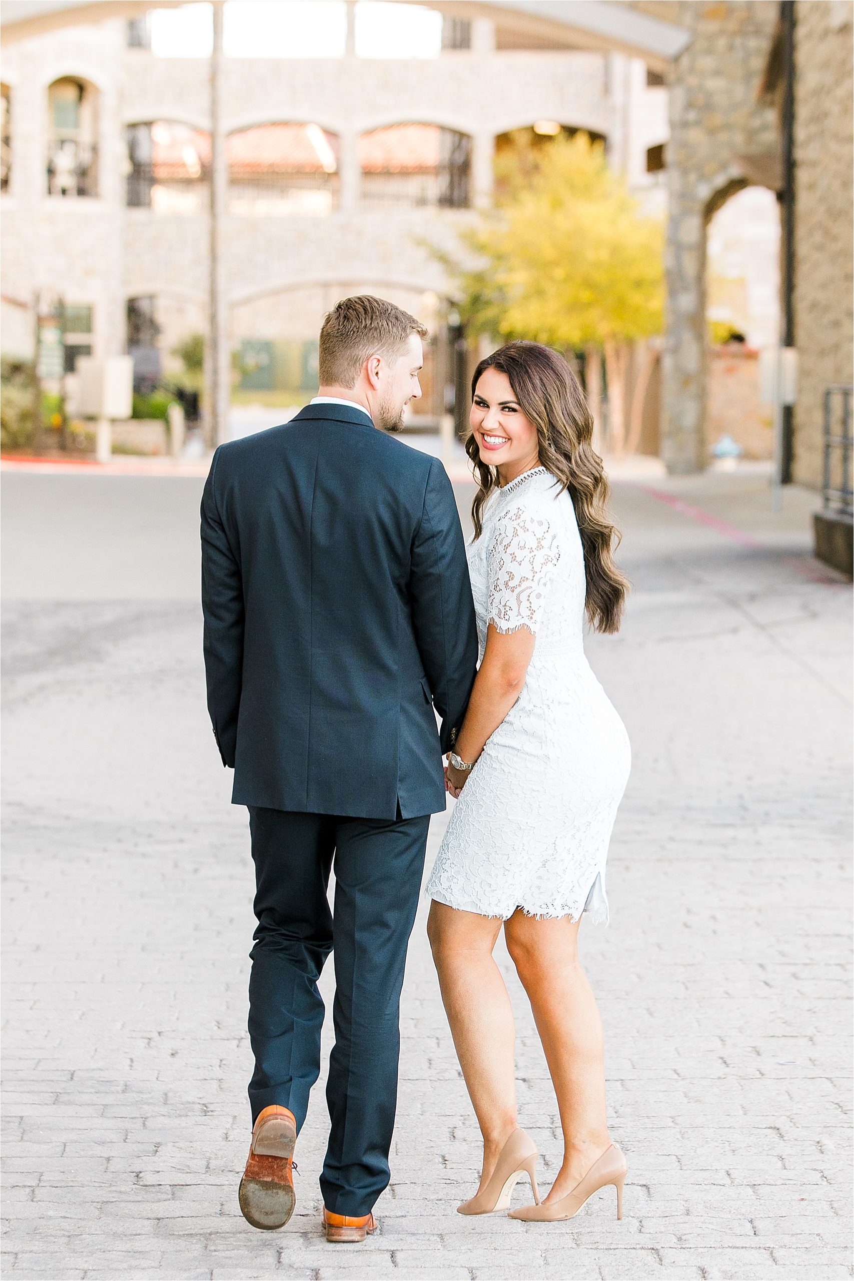 A bride to be holds hands with her fiance as they walk away and she smiles back at the camera while he looks at her during their engagement session at adriatica village in McKinney, Texas 