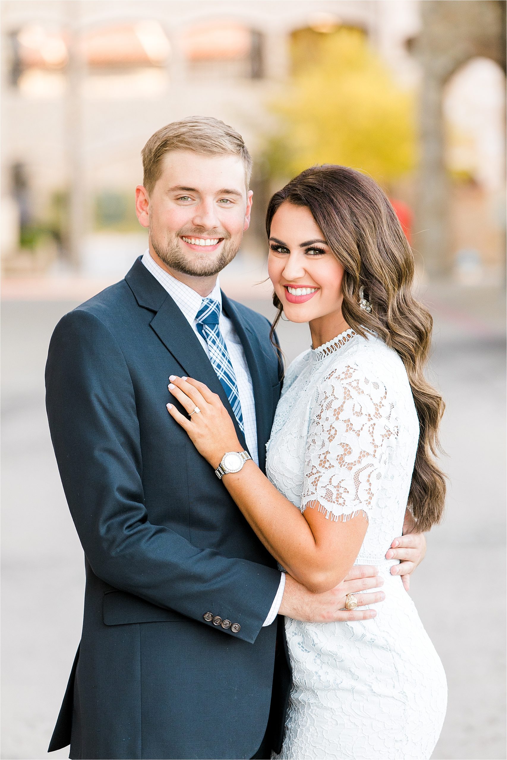 An engaged couple in a blue suit and a lace dress smile at the camera during their engagement session at adriatica village in McKinney, Texas 