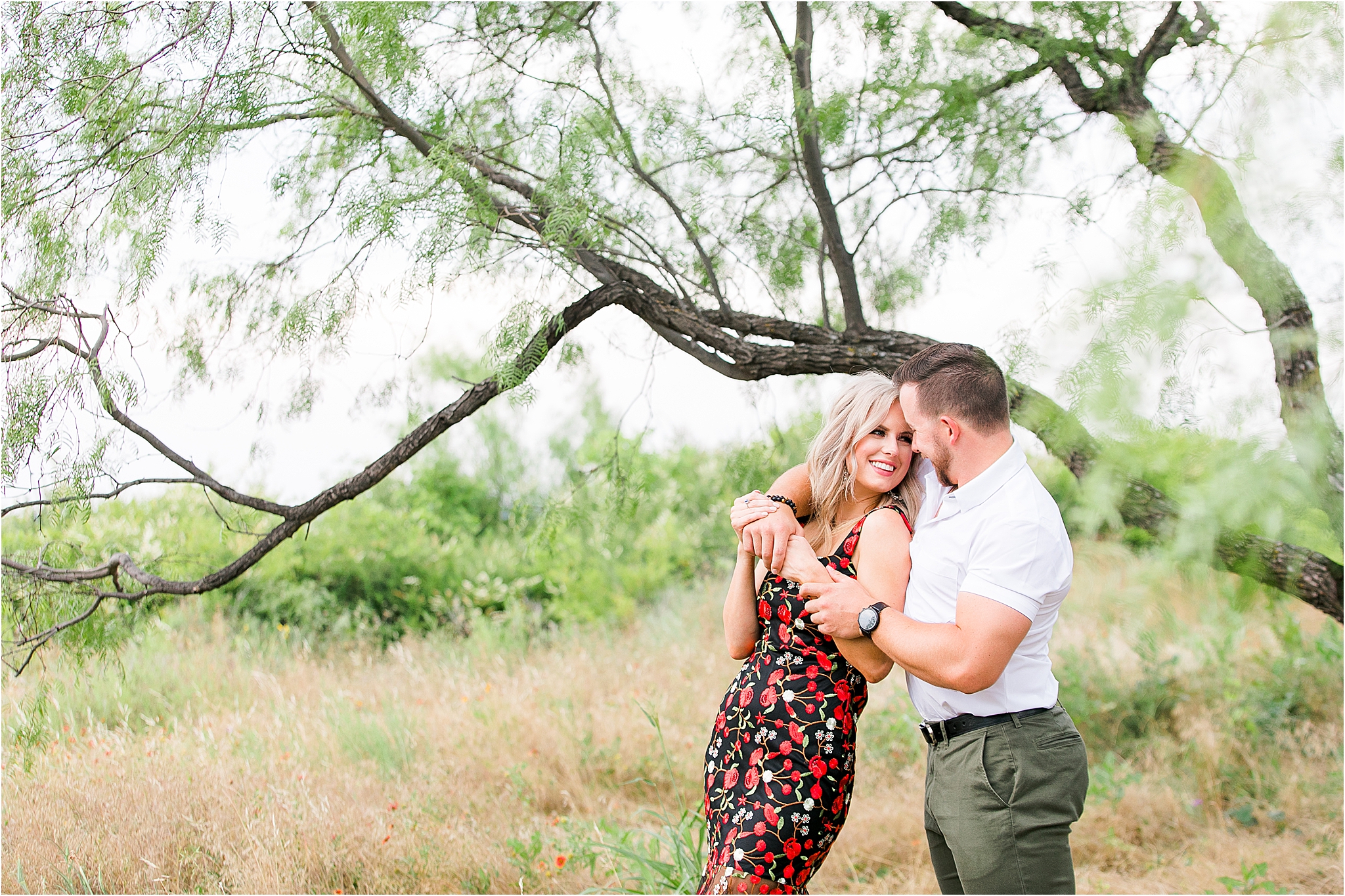 A bride to be glances back at her groom as he hugs her from behind underneath a tree at Tandy Hills Nature Preserve during their Fort Worth Engagement Session 
