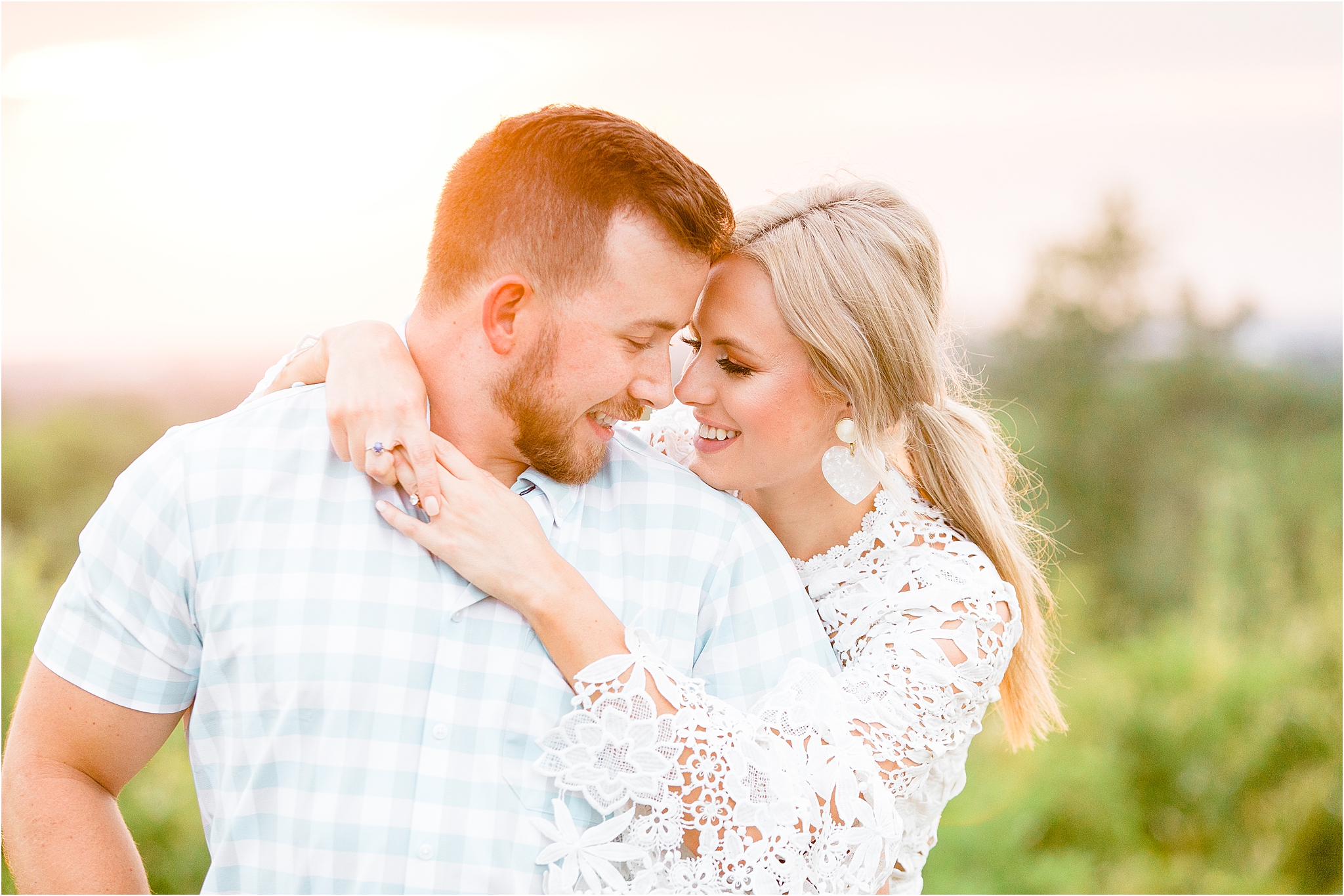 A blond dallas bride to be hugs over her fiance in a blue and white plaid shirt in front of a vibrant sunset in Fort Worth, Texas during their engagement session 