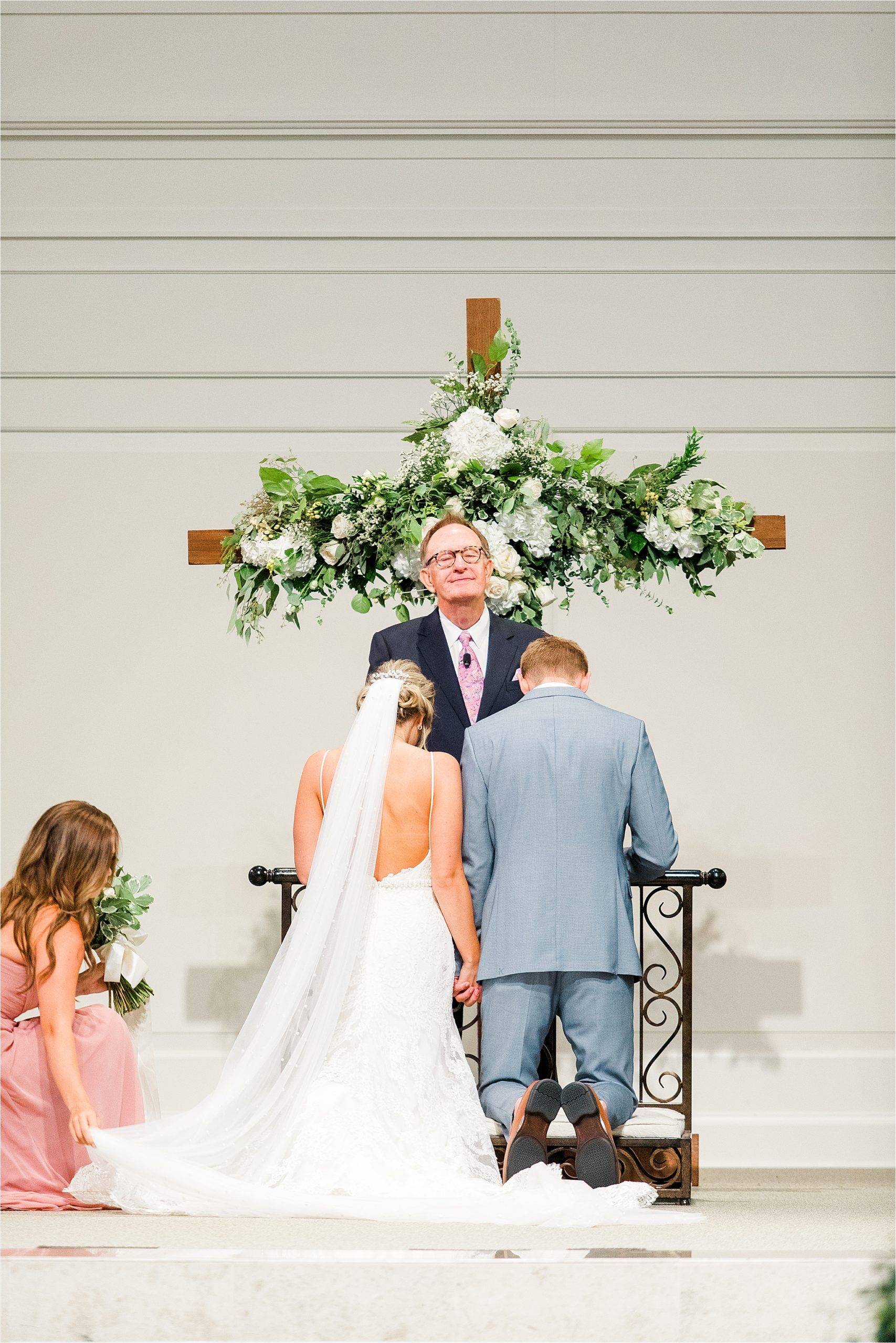 A bride and groom kneel before the cross as her bridesmaid adjusts her train during her prestonwood chapel ceremony in Plano, Texas 