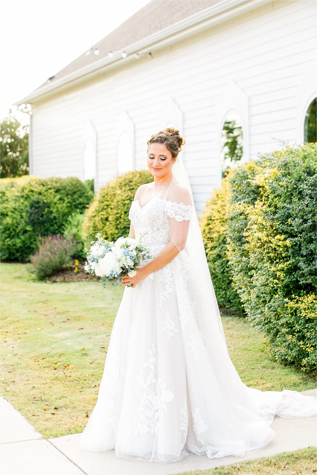 A bride softly smiles and looks down while posing for a bridal portrait in front of a white chapel at Rustic Grace Estate 
