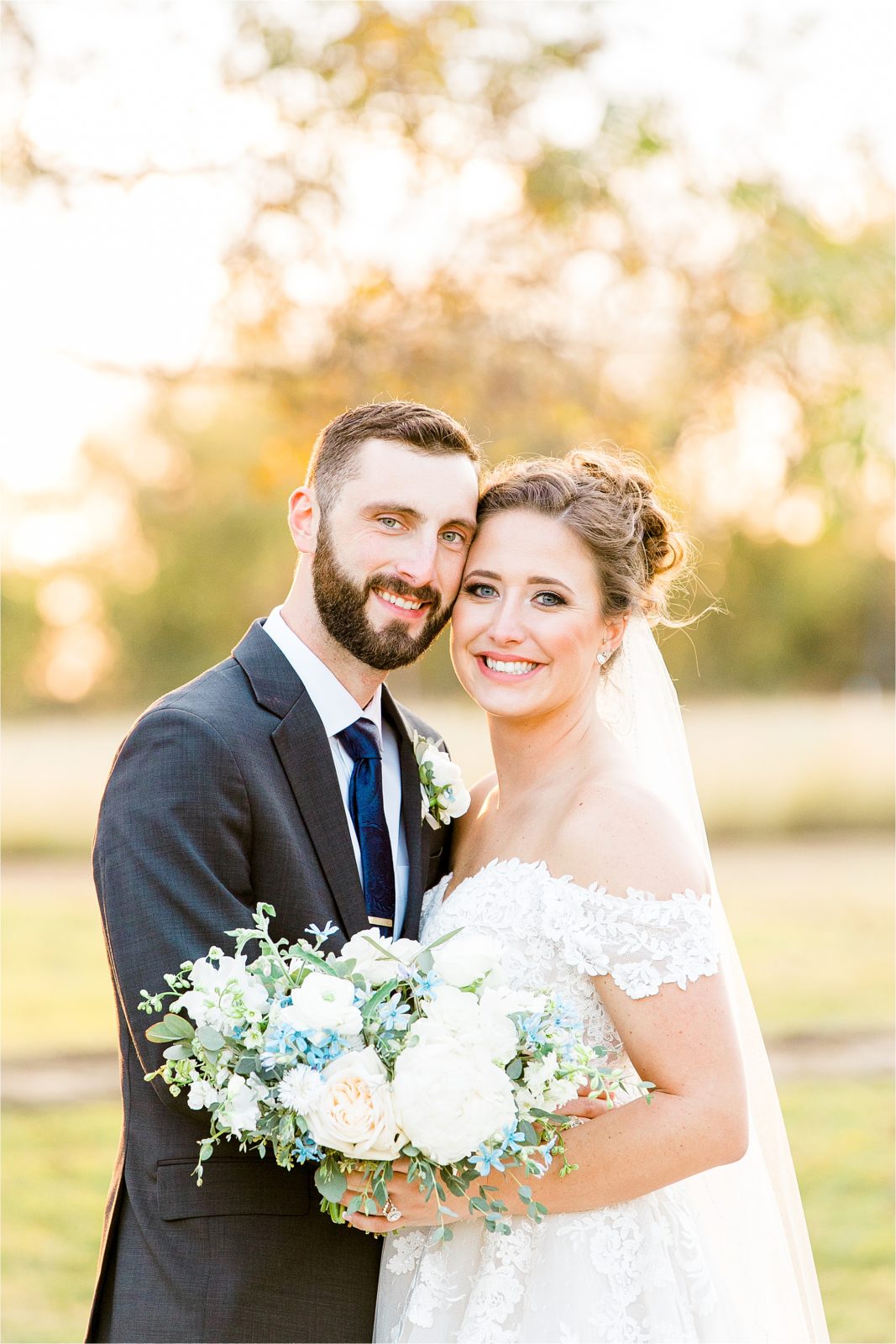 A newly wed couple happily smiles at the camera cheek to cheek holding her white and blue flowered bouquet on a sunny, fall wedding day at Rustic Grace Estate 