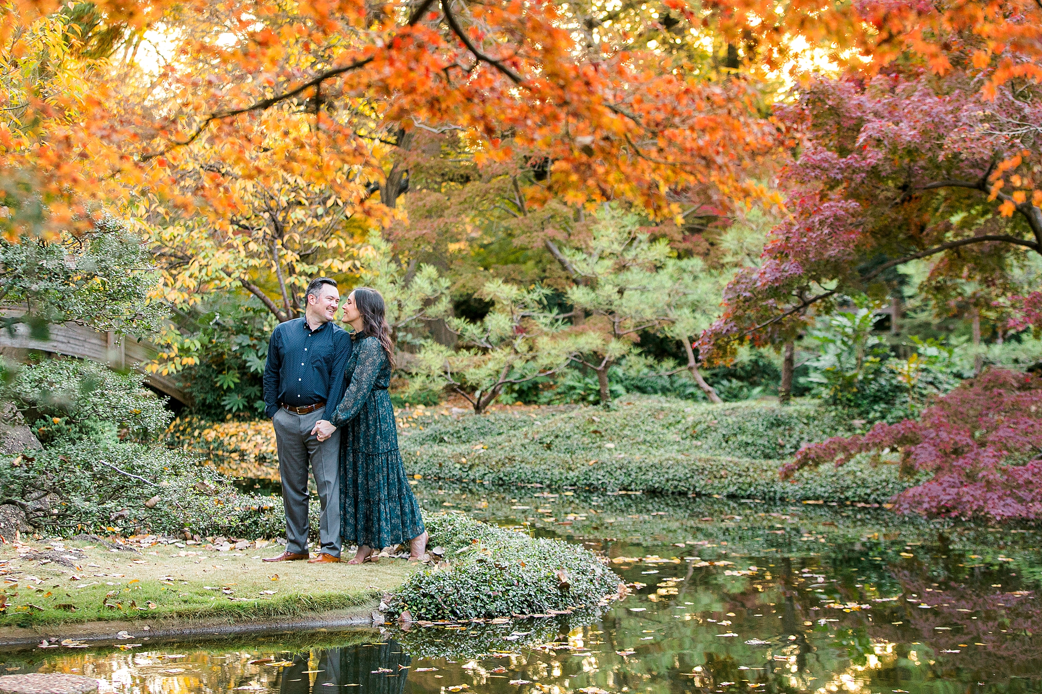 An engaged couple nuzzles under a tree full of orange leaves inside the Japanese Gardens at The Fort Worth Botanic Garden