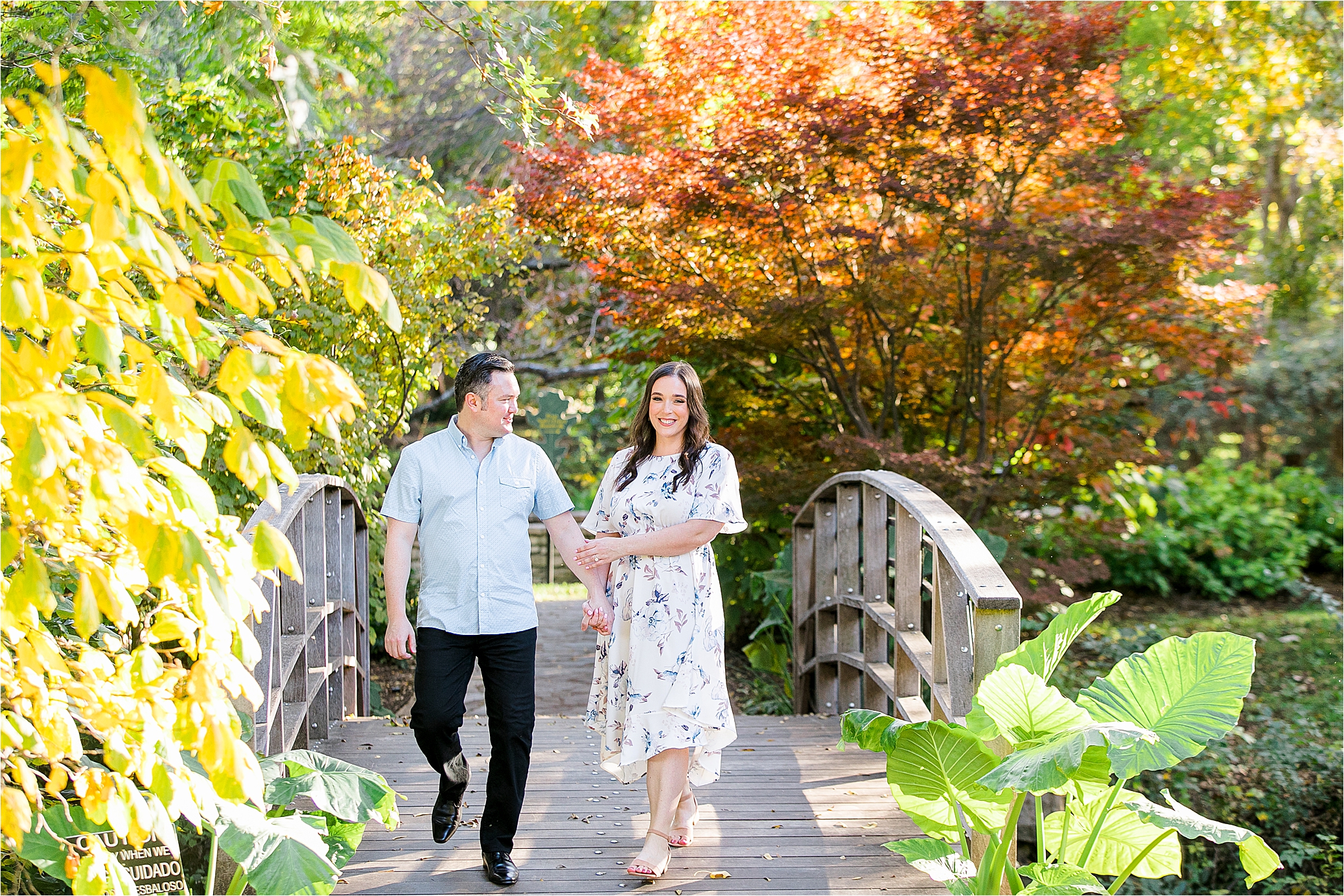 A couple holds hands and walks over a wooden bridge on a sunny day their For Worth Engagement Session at The Botanic Garden