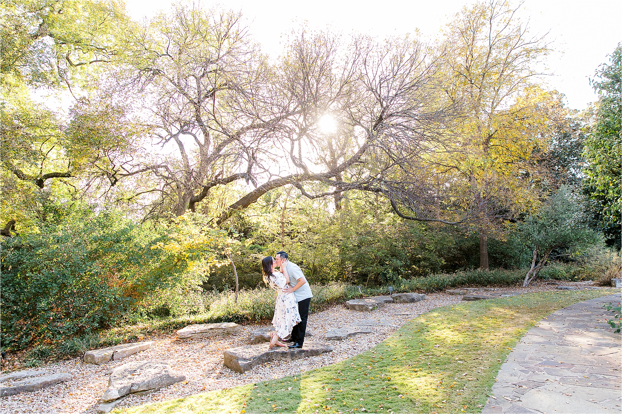 A couple dips and shares a kiss in front of some trees during their engagement session on a warm, sunny day in Fort Worth Texas
