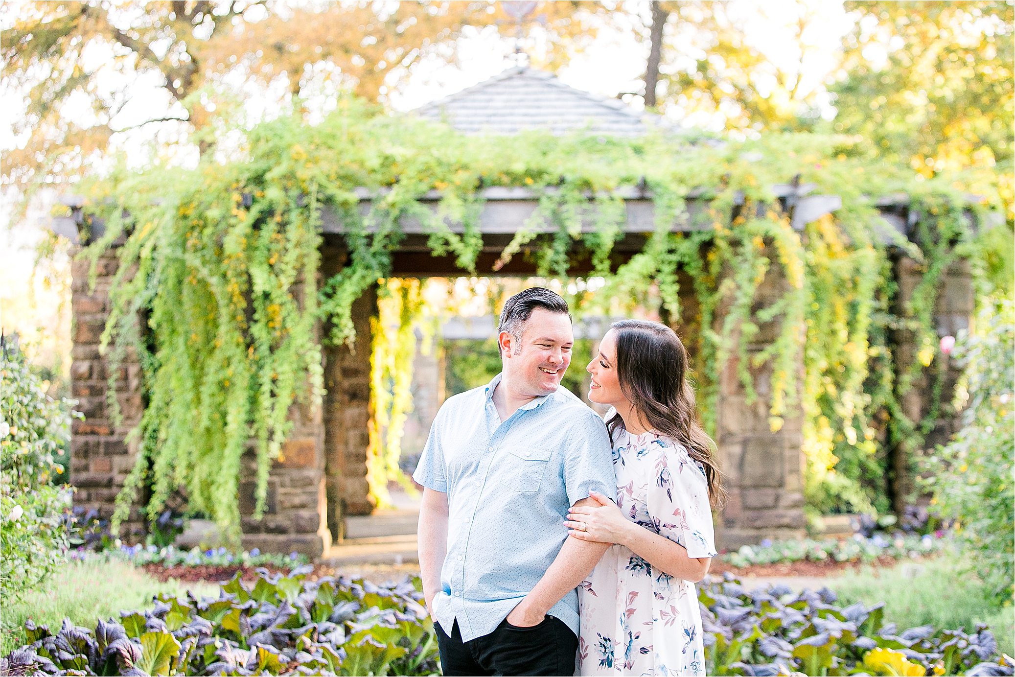 A bride to be hugs her groom from behind in front of an arch of greenery on a sunny, fall day at The Fort Worth Botanic Garden during their engagement session 