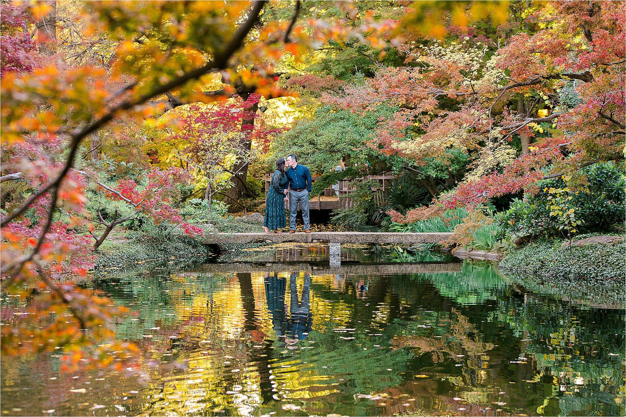 A couple shares a kiss on a bridge reflecting in the pond below and underneath colorful leaves during their Fall Engagement Session at The Fort Worth Botanic Garden 