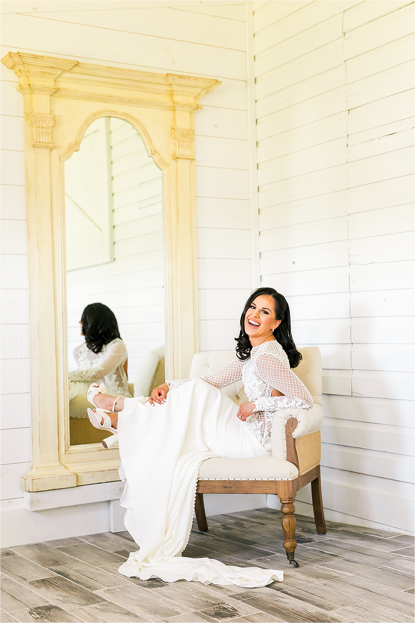 A bride laughs toward the camera while sitting in a chair in her all-white bridal suite 