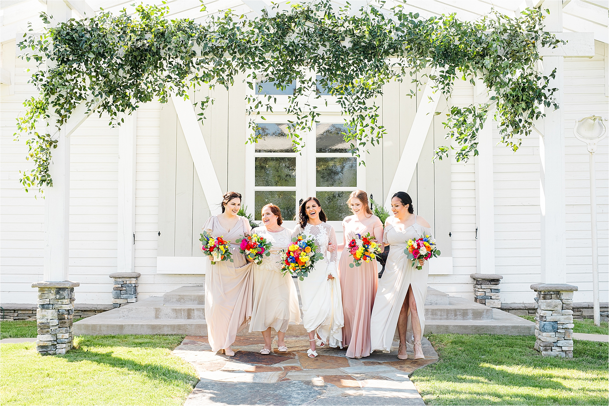 A joyful bride and her bridesmaids laugh while walking together in front of her white wedding venue in East Texas 