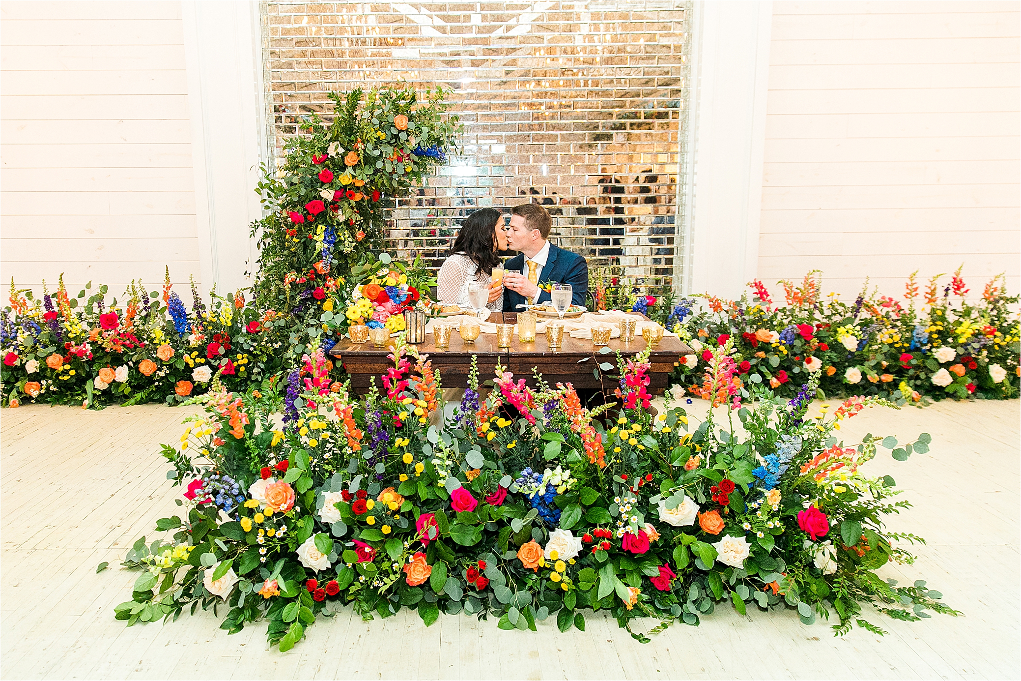 A newly married couple kisses at their head table surround by colorful flowers on their wedding day at Pineway Farms