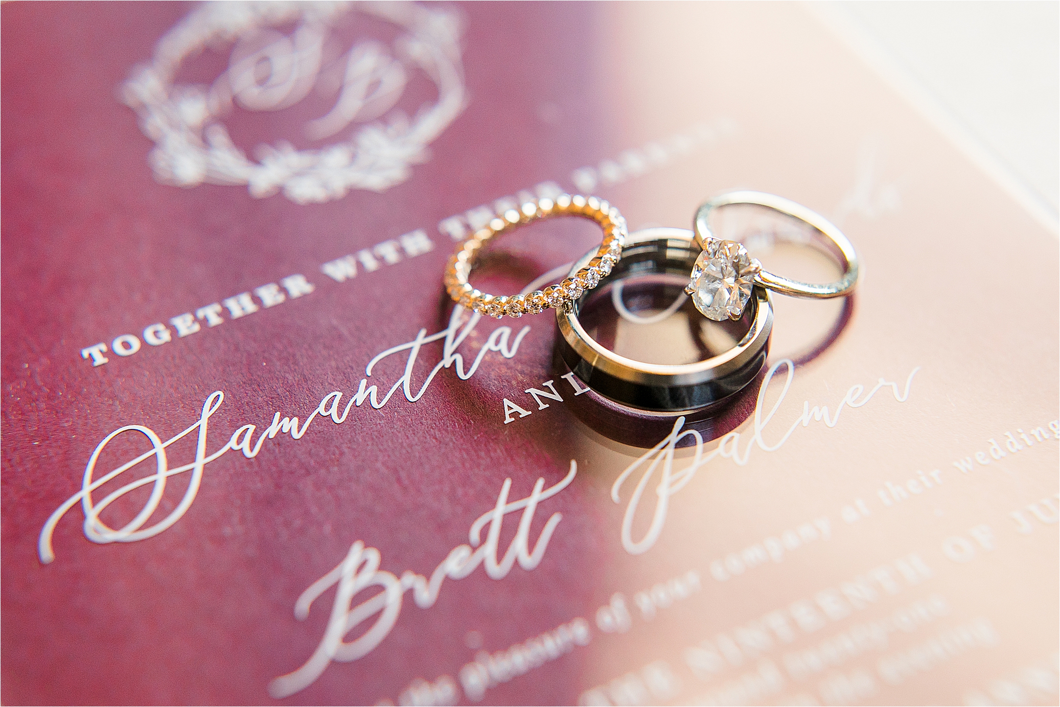 wedding Rings displayed over a maroon acrylic invitation that reads Samantha & Brett by Hill Country Wedding Photographer Jillian Hogan 