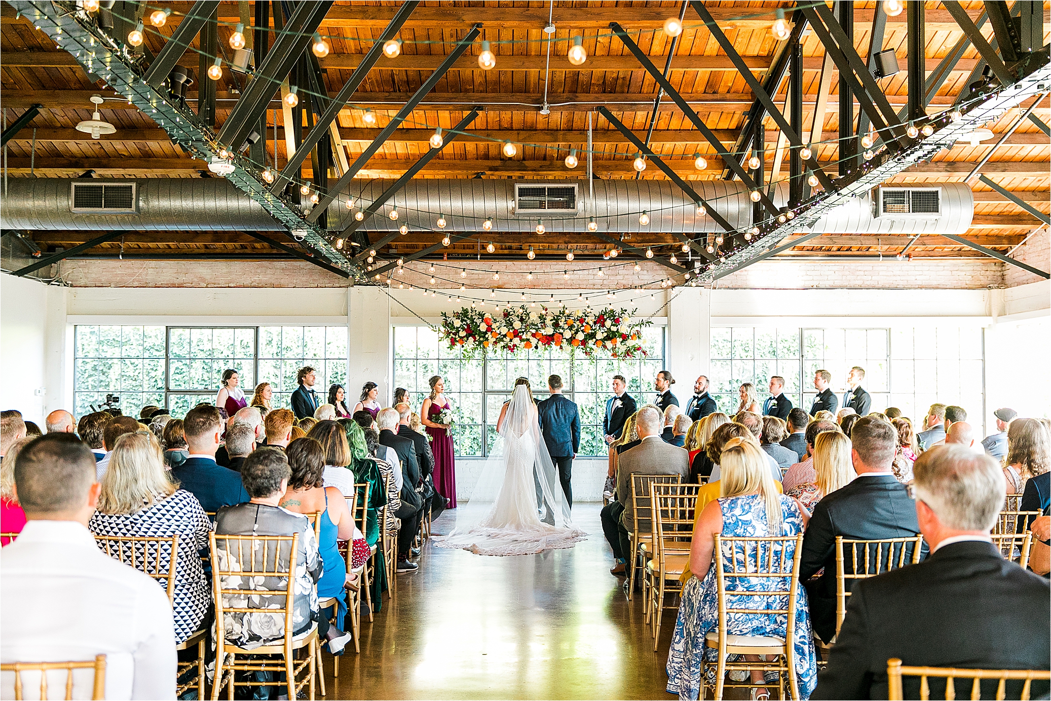 A wide view of a ceremony site with a hanging floral arch and twinkle lights and the back of the bride and groom at Hickory Street Annex Wedding in Dallas, Texas with Wedding Photographer Jillian Hogan Photographer
