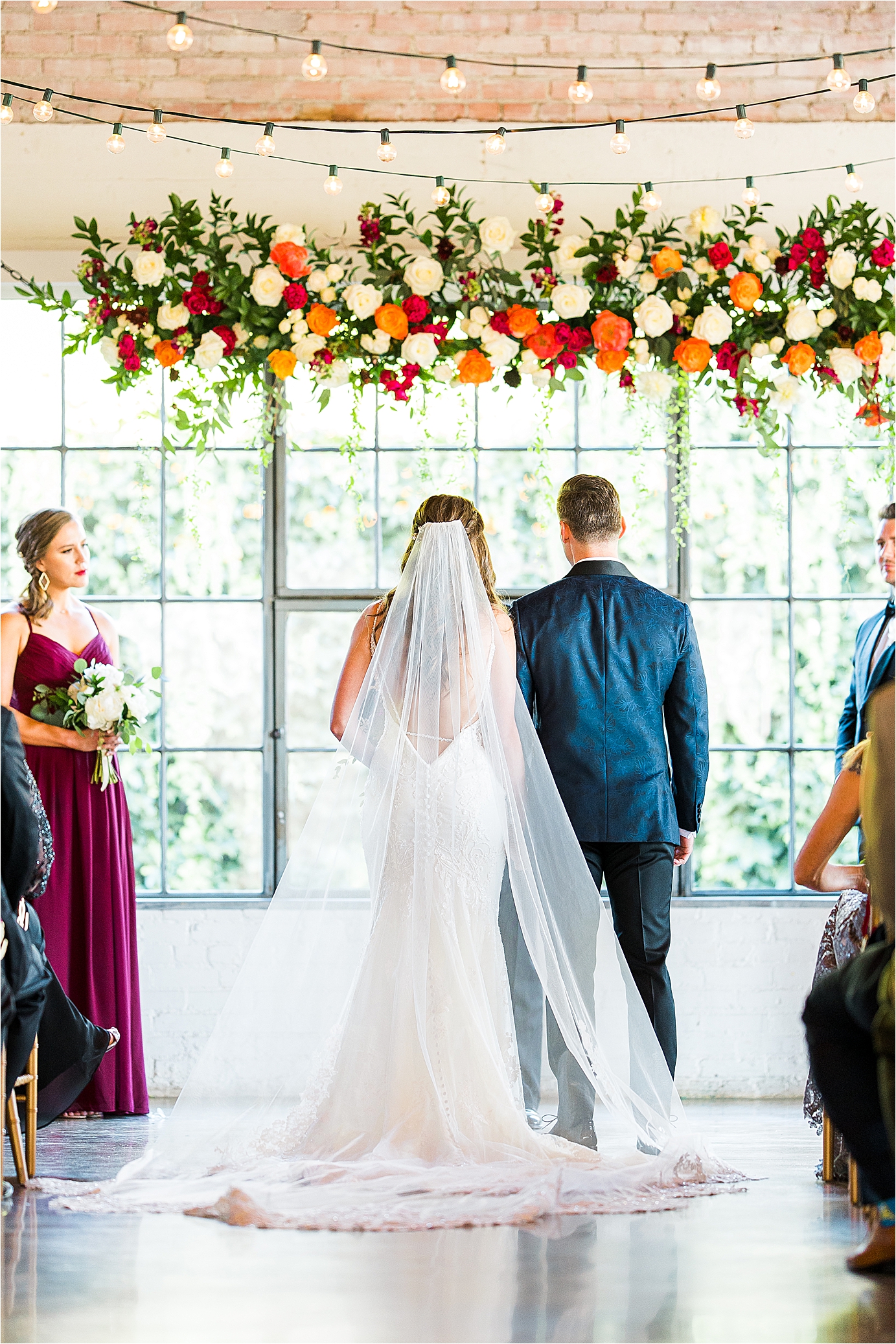 A bride and groom hold hands as you get a view of the back of the bride's dress and veil during her floral filled ceremony at Hickory Street Annex in Dallas, Texas 