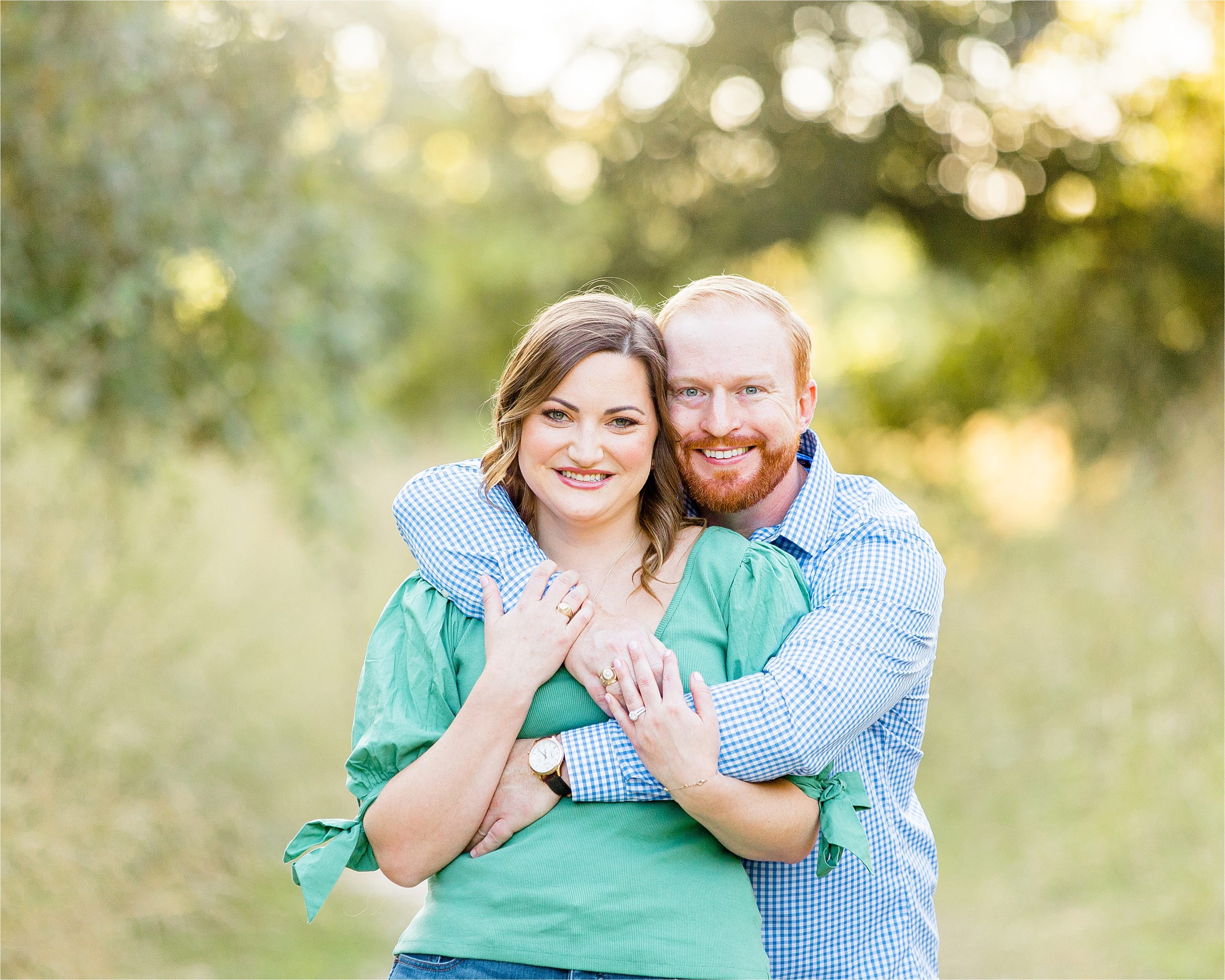 A guy in a blue shirt hugs over his fiance while they smile big at the camera during their sunset engagement session in Boerne, Texas