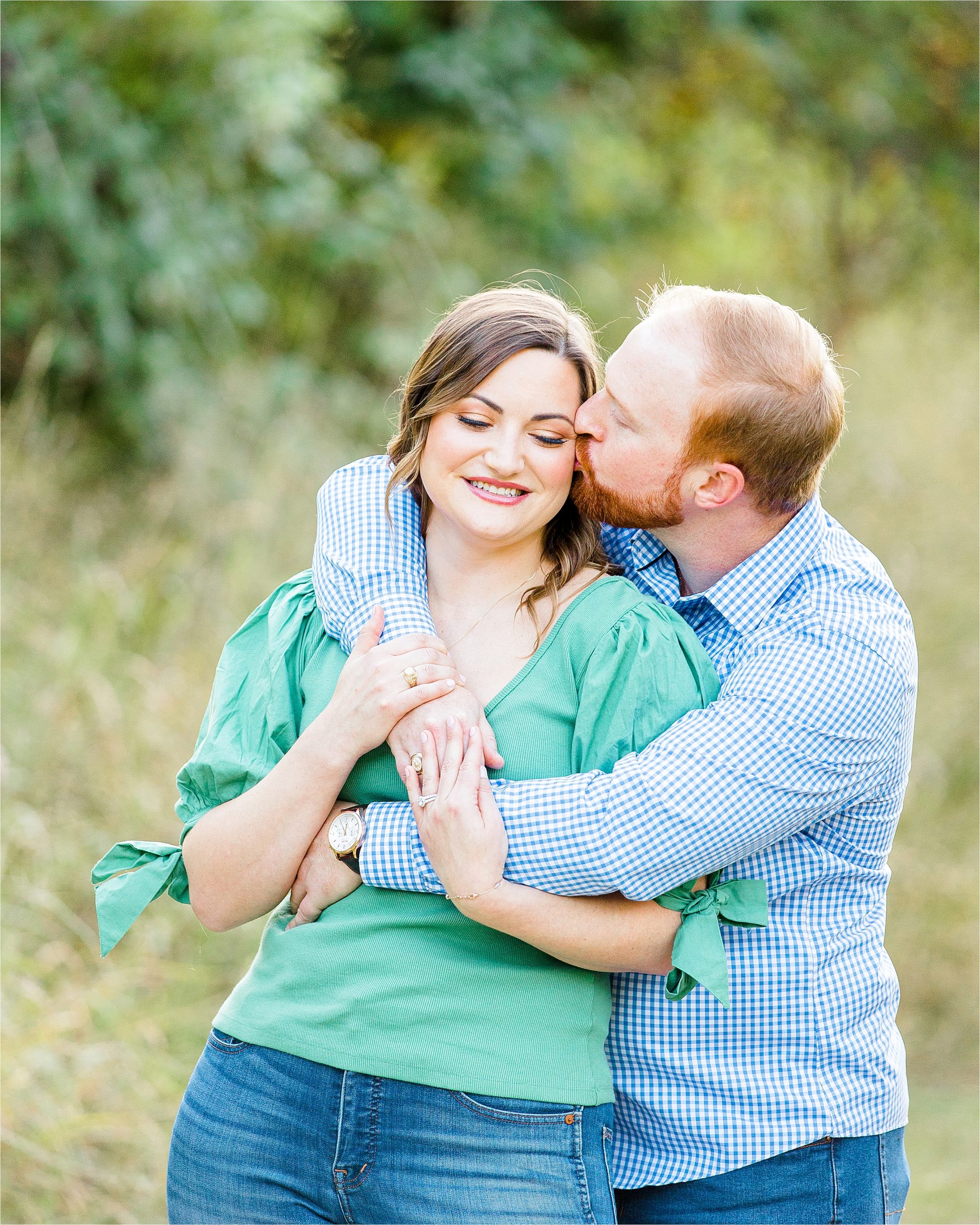 A groom to be hugs over his fiance's shoulder and gives her a big kiss on the cheek as she smiles during their outdoor engagement session at Cibolo Creek Nature Center. 