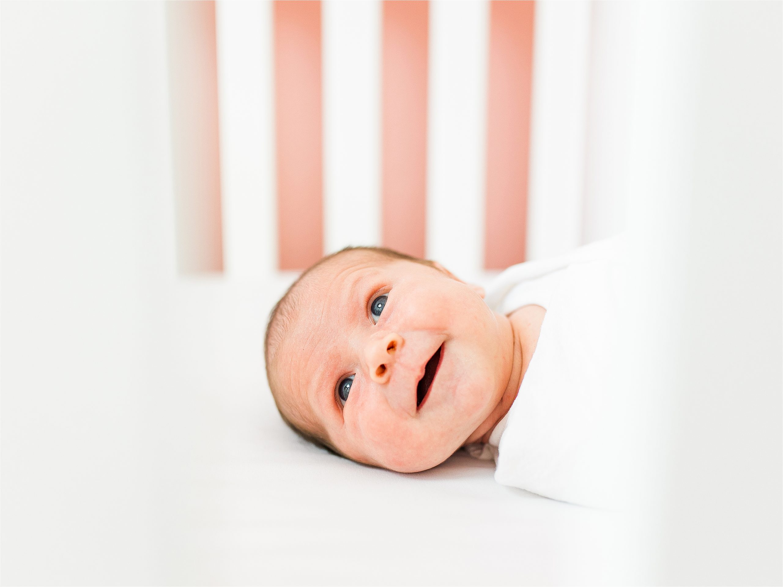 A newborn baby girl smiles in her crib swaddle in white during her in home newborn portraits in San Antonio, Texas