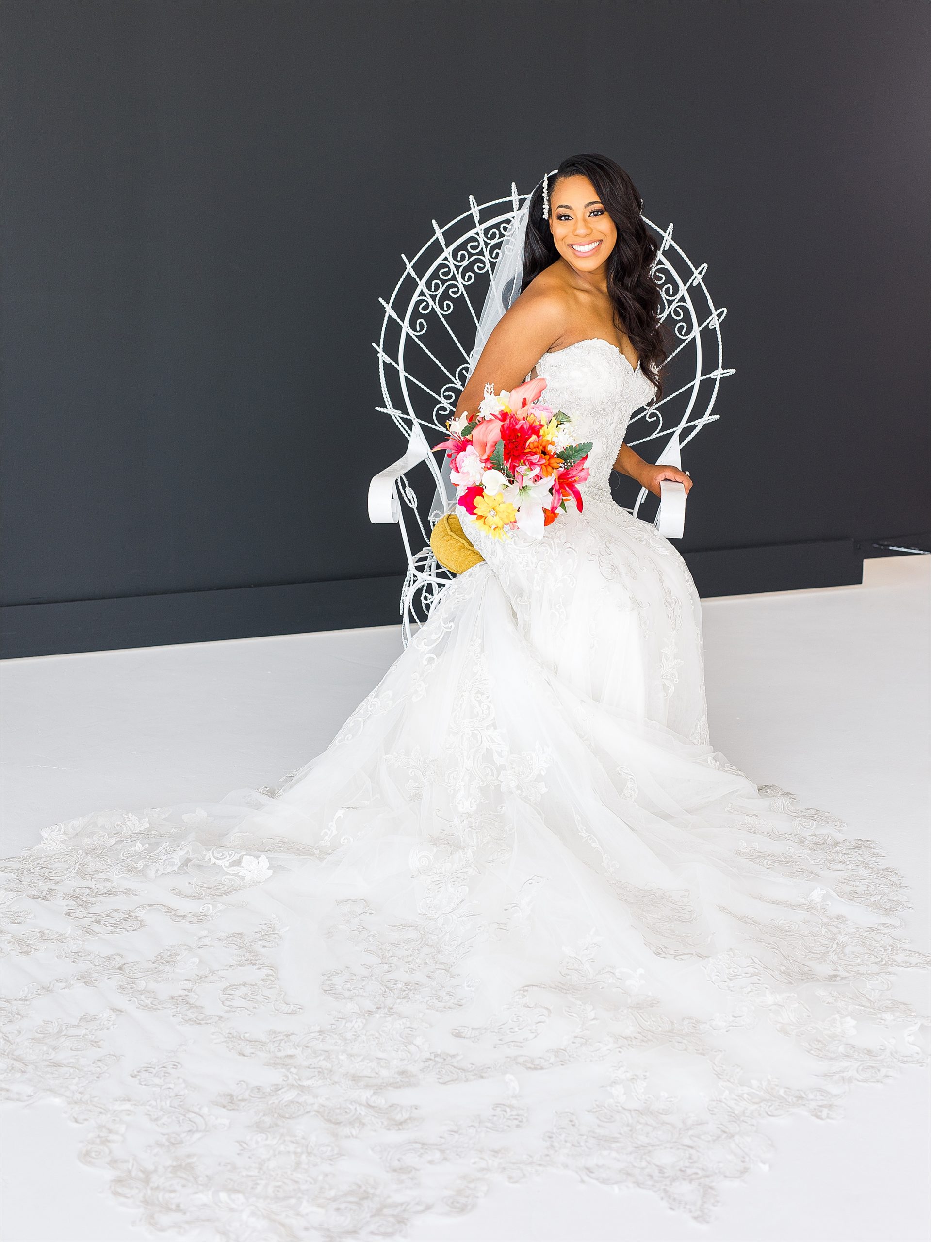 A bride sits in a white game of thrones looking chair with a colorful bouquet and her lace dress covering the floor during her San Antonio Studio Bridal Portraits 