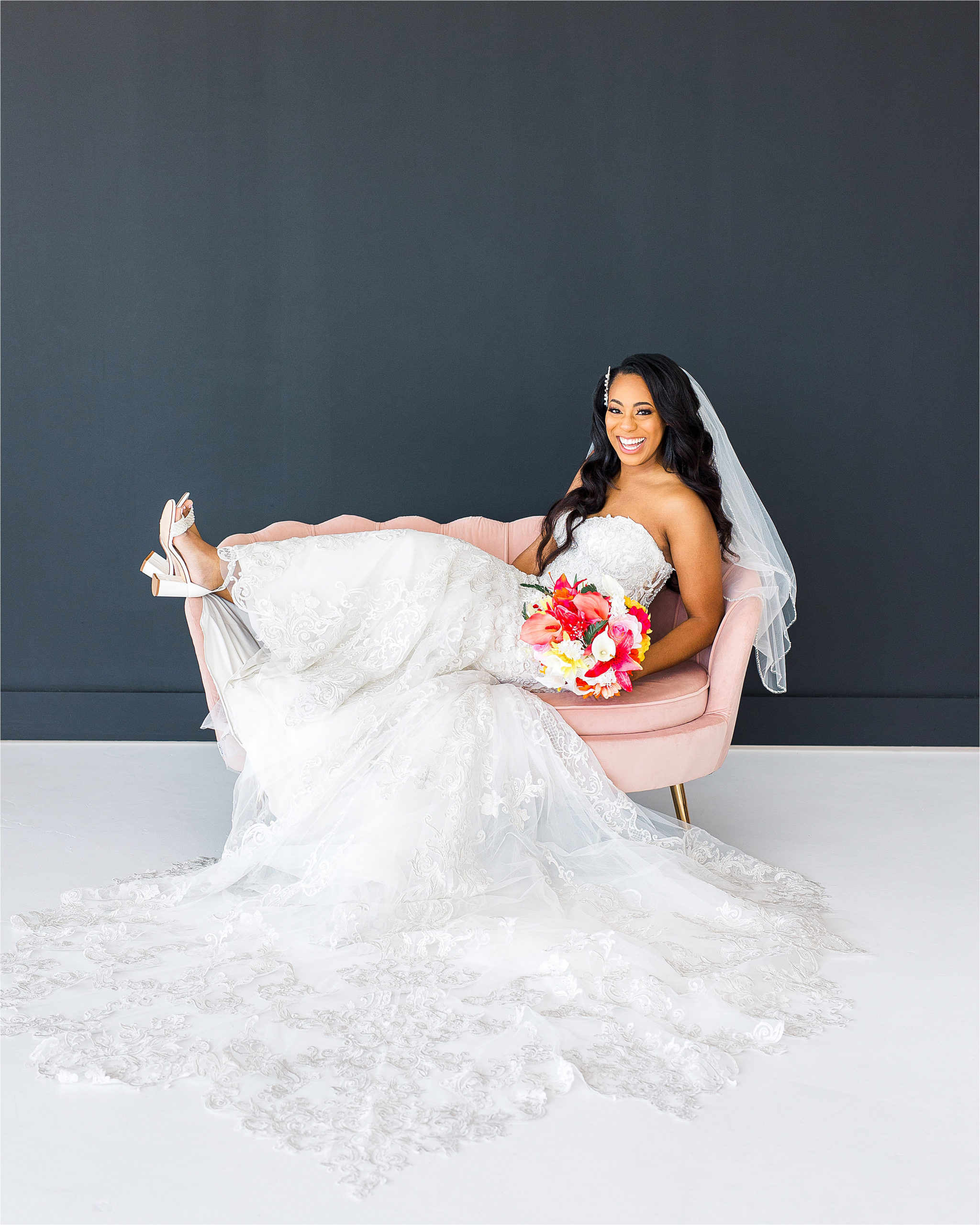 A bride laughs really hard sitting on a pink couch in front of a black backdrop with her lace wedding dress spread out during her studio bridal sessions in San Antonio, Texas 