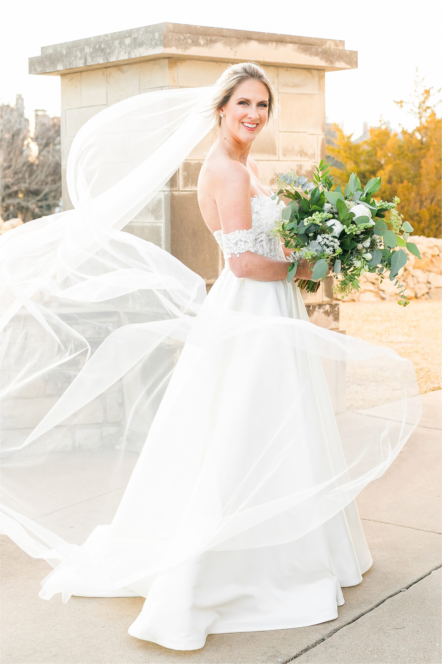 A bride smiles at the camera with a lush bouquet and the sunset behind her as her veil blows in the wind circling around her during her adriatica village bridal portraits in McKinney, Texas with Hill Country Wedding Photographer Jillian Hogan 