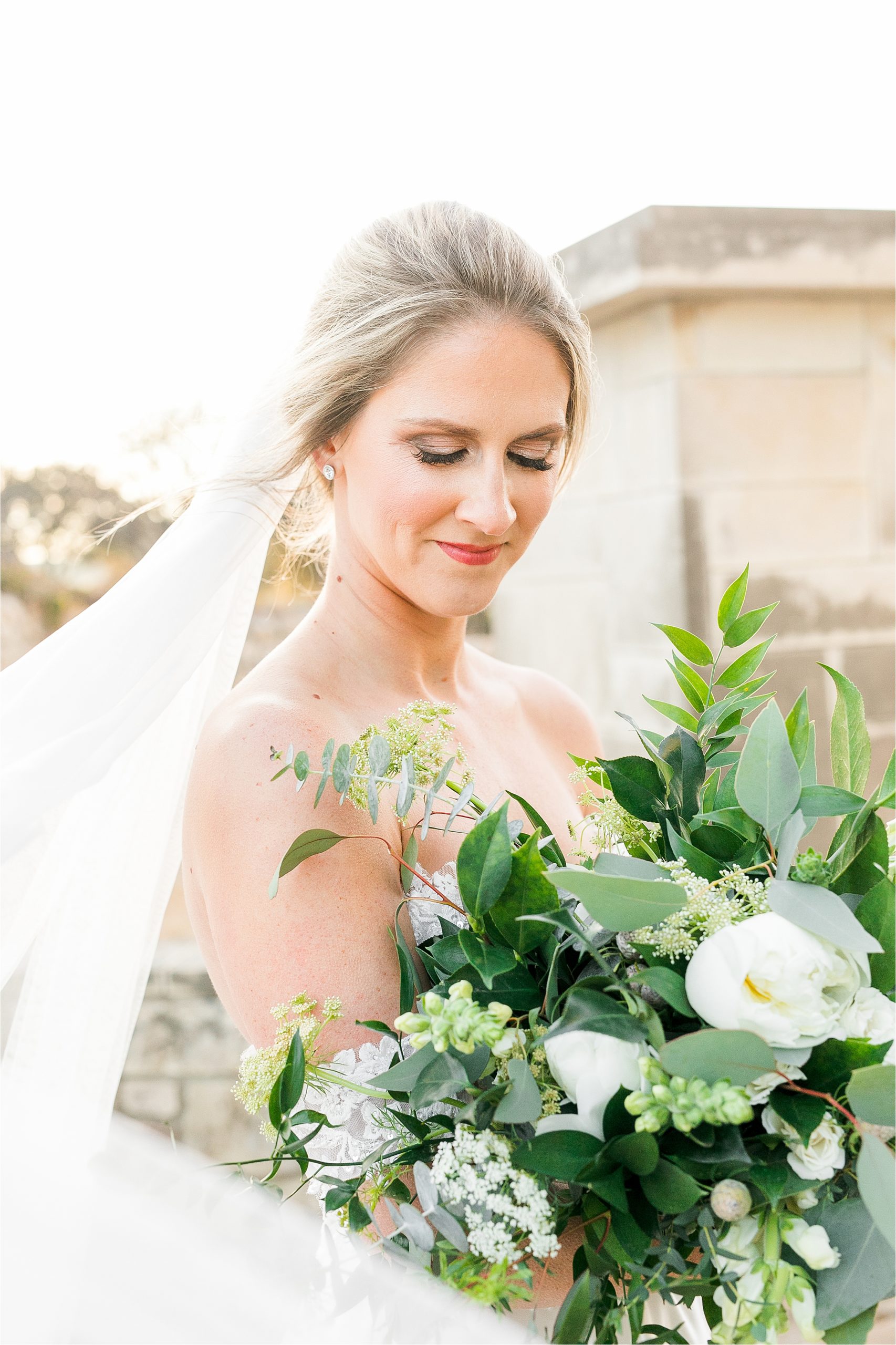 A bride with her hair pulled back shares a soft smile and looks down at her flowers with a veil behind during her warm, winter bridal portraits at adriatica village in McKinney, Texas with Austin Wedding Photographer Jilian Hogan 