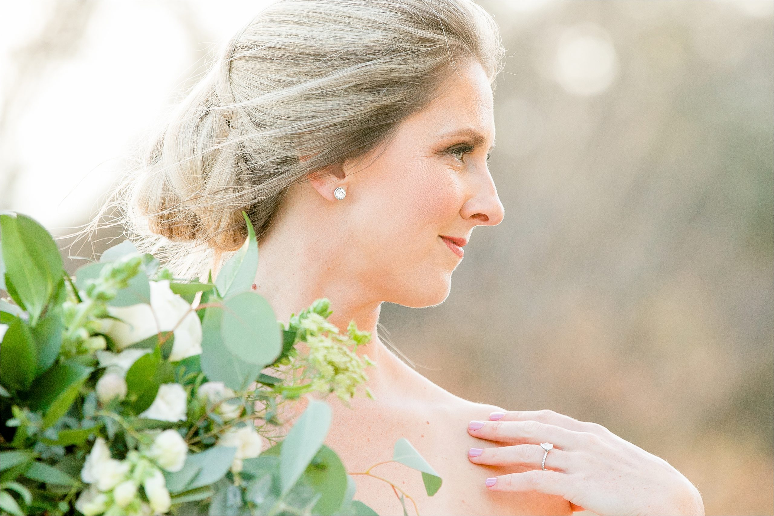 A closeup of a bride looking off in the distance with her engagement ring and greenery filled bouquet with the sunset behind her. 