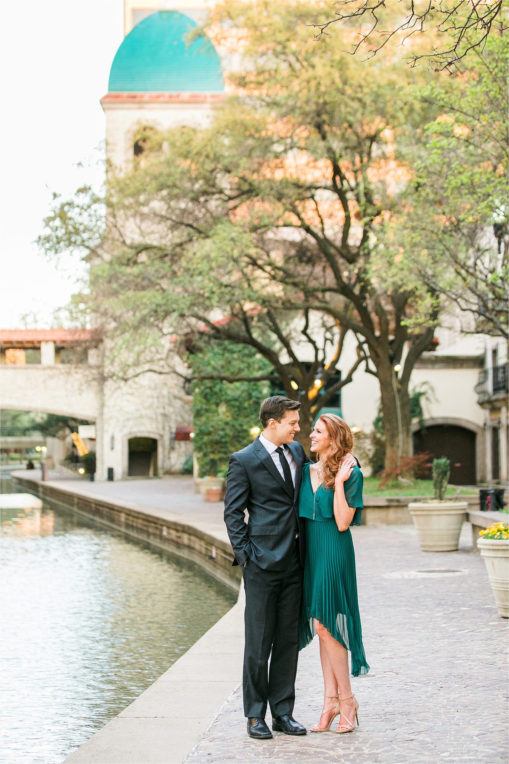 A groom dressed in a suite hugs over the shoulder of his Miss Texas 2016 Bride while she wears an emerald dress for their riverwalk inspired engagement session with San Antonio Wedding Photographer Jillian Hogan Photography 