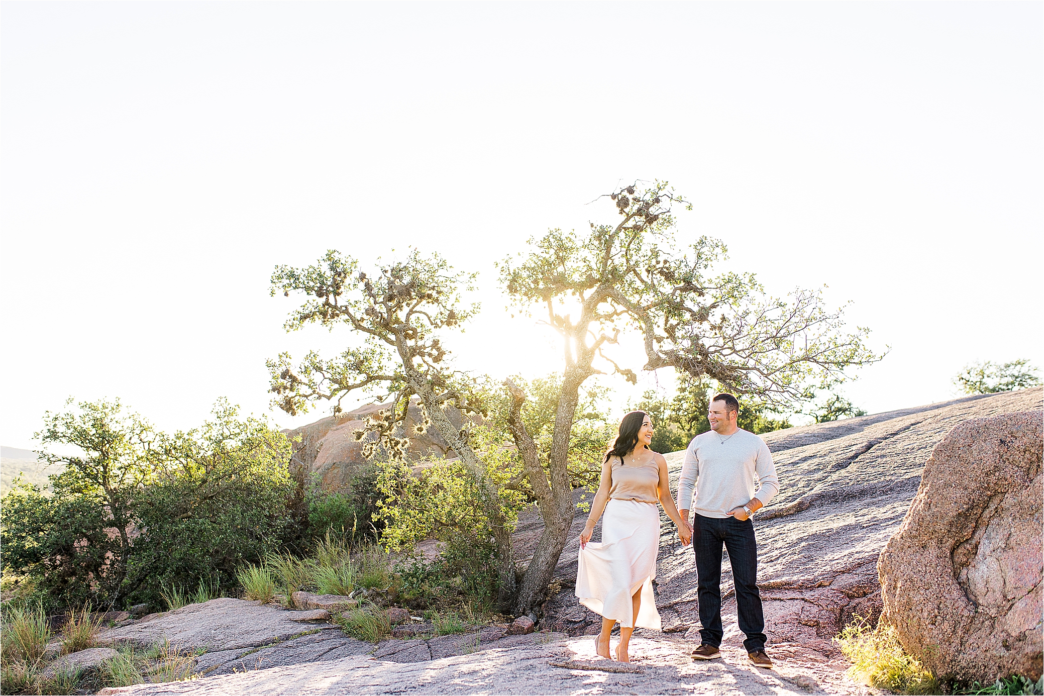 An engaged couple holds hands in front of a tree that is blocking the sun as the look at each other during their Texas Hill Country Engagement Session 
