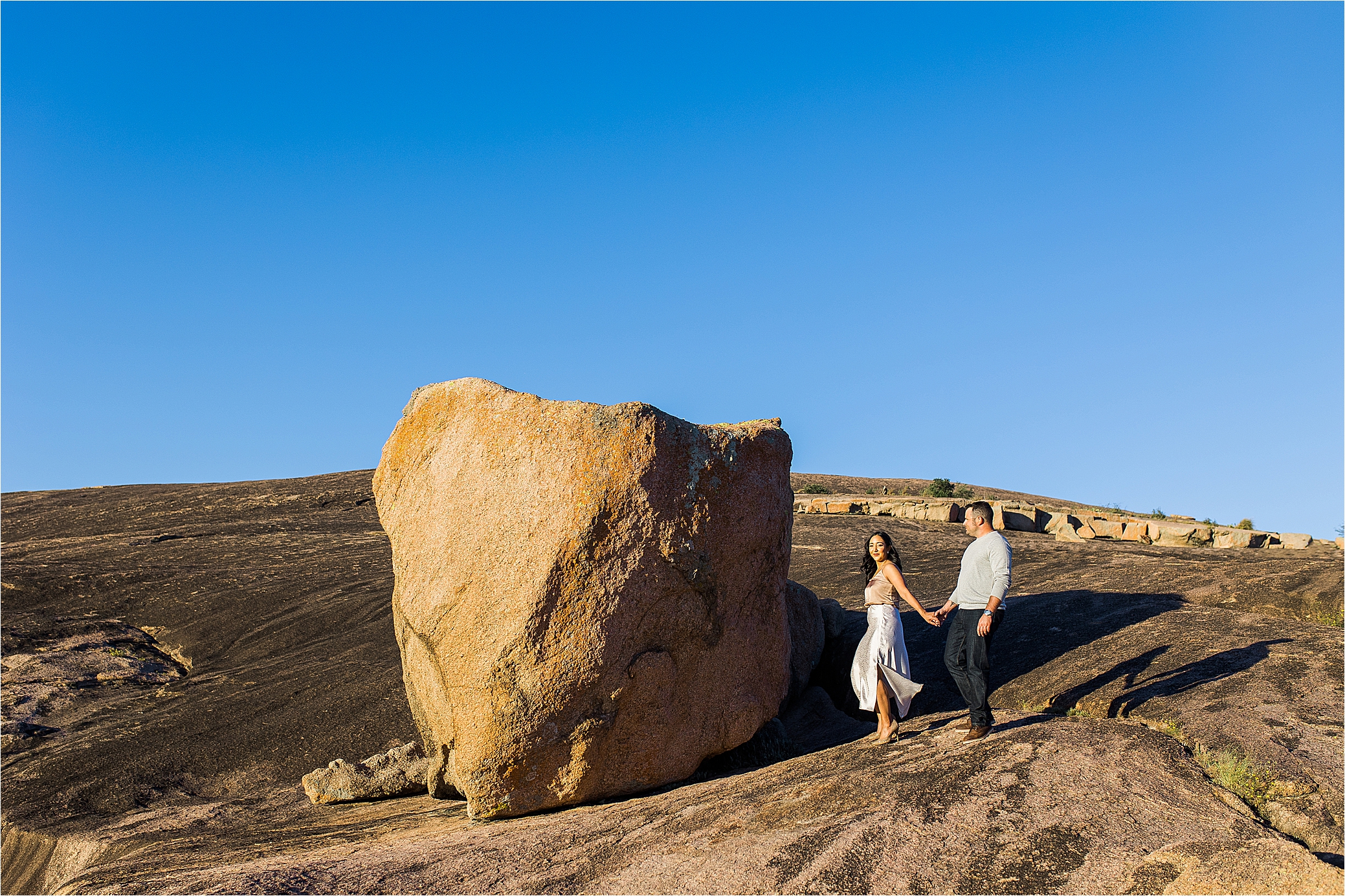 A woman in a white long skirt pulls her fiance as they walk in front of a large boulder in the sun at Enchanted Rock State Park 
