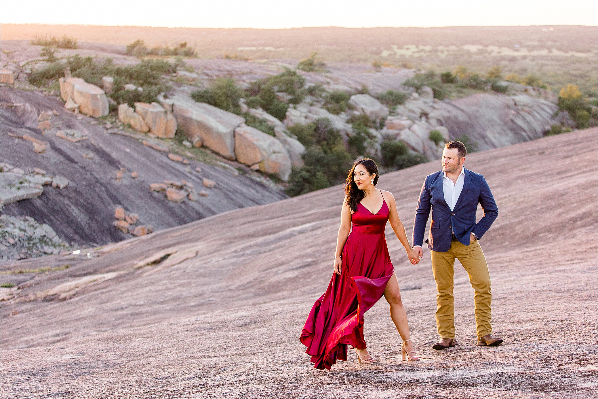 A couple holds hands and looks off left in the distance as her red dress blows in the wind at Enchanted Rock State Park during their Texas Hill Country Engagement Session 