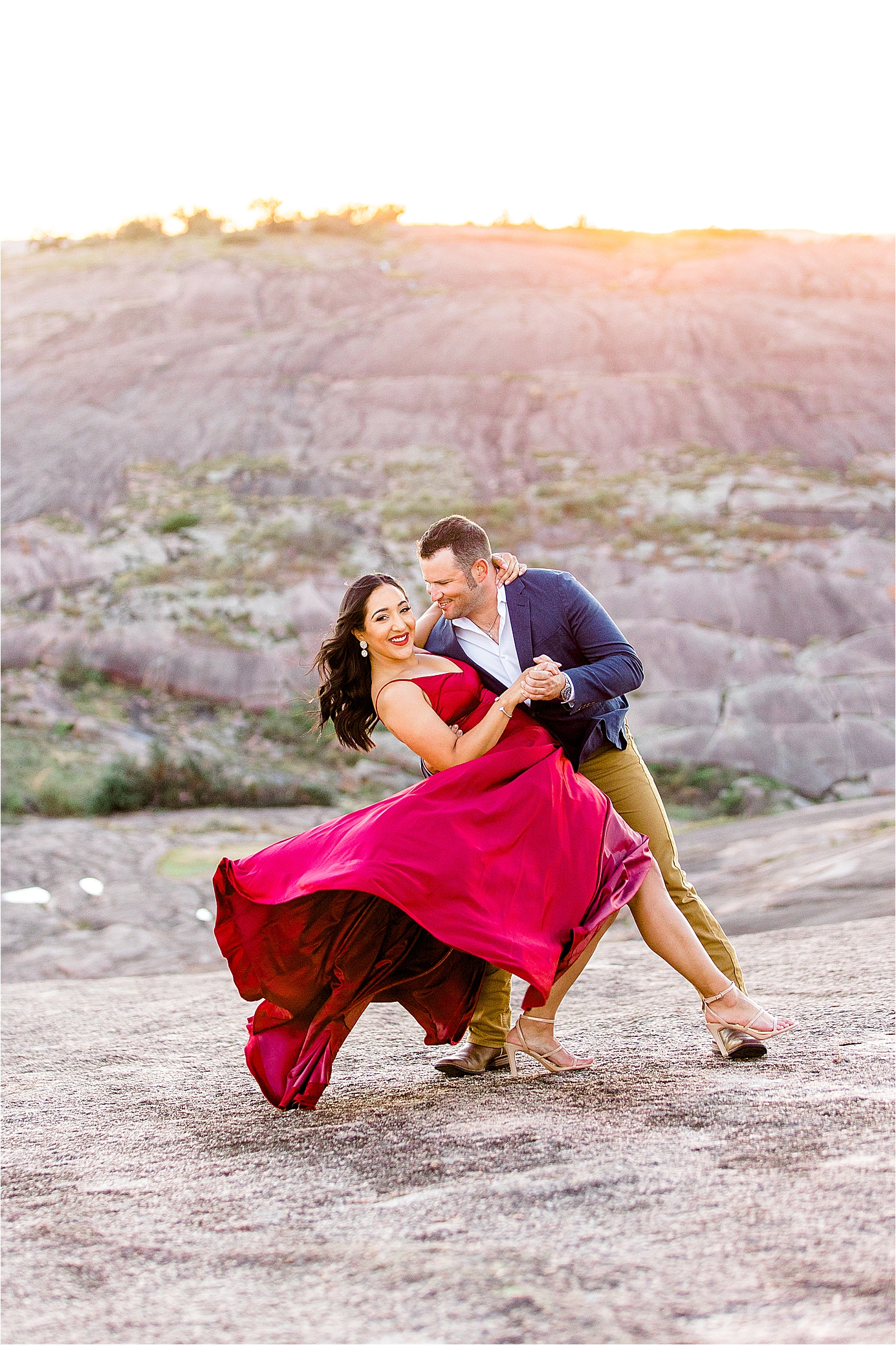 A couple dips and the girl smiles at the camera as her red dress blows in the wind during their Texas Hill Country Engagement Session at Enchanted Rock
