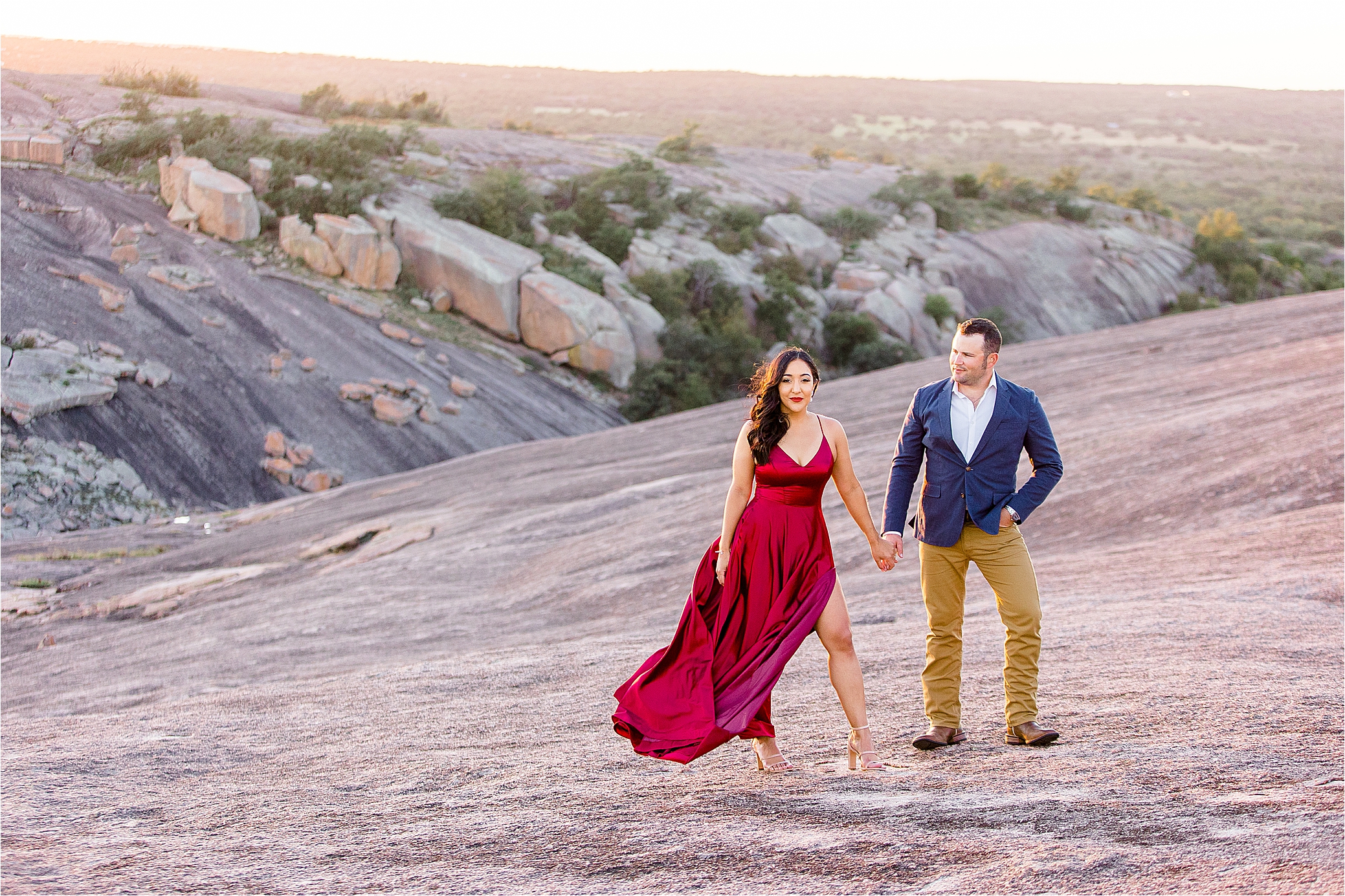 A girl stares at the camera as her fiance. holds her hand and gazes at her. Her red dress blows in the wind at the top of Enchanted Rock with Hill Country Engagement Photographer Jillian Hogan 