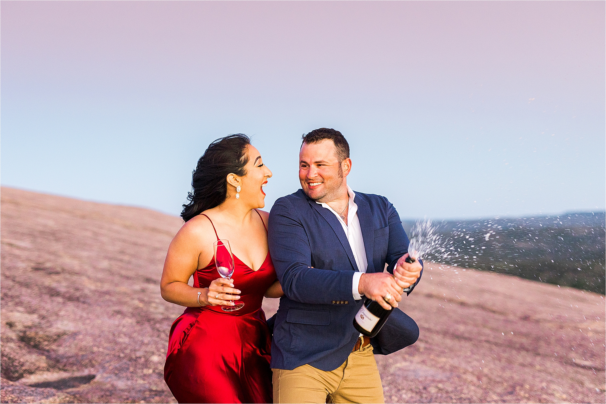 An engaged couple laughs at each other and sprays champagne in front of a purple sunset at Enchanted Rock in the Texas Hill Country for their engagement session
