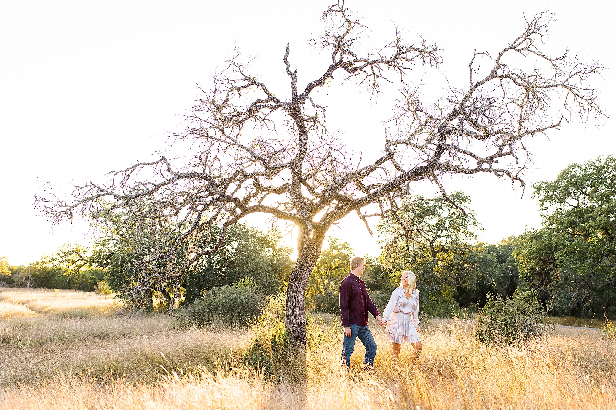 A blonde bride to be pulls her groom along in a maroon shirt under a tree in a field with the sun shining behind them for their Texas Hill Country Engagement Session with Wedding Photographer Jillian Hogan 