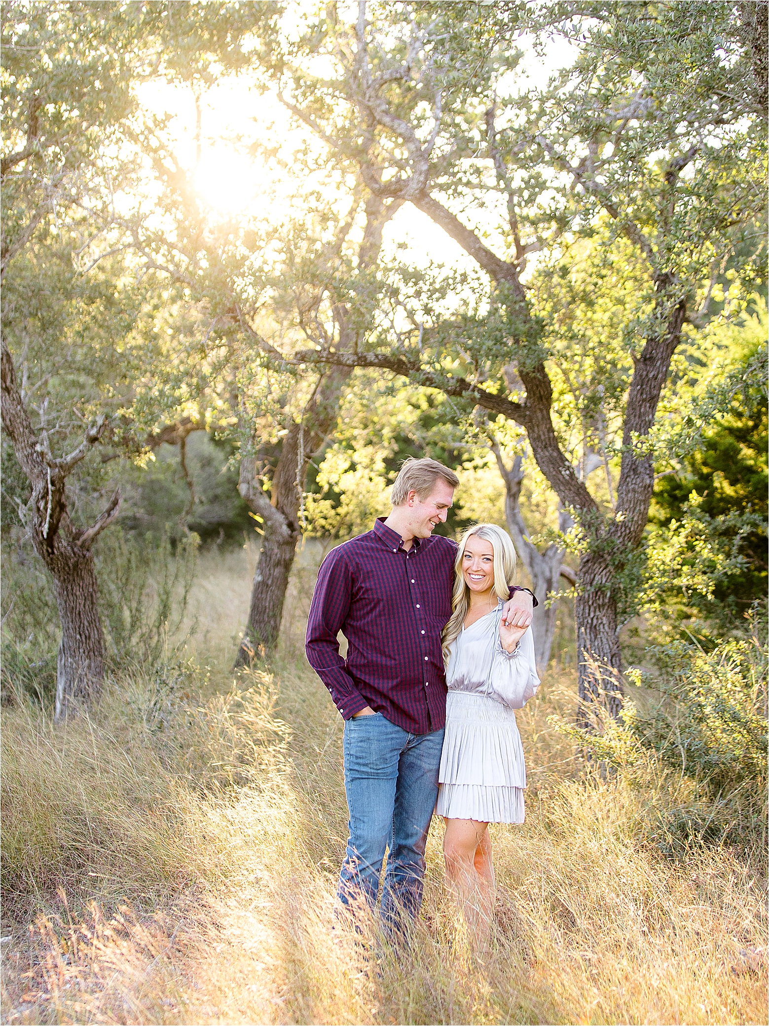 A couple poses in the field with trees and sunshine behind them as the guy puts his arm around her for their Fall Hill Country Engagement Session with San Antonio Wedding Photographer Jillian Hogan 