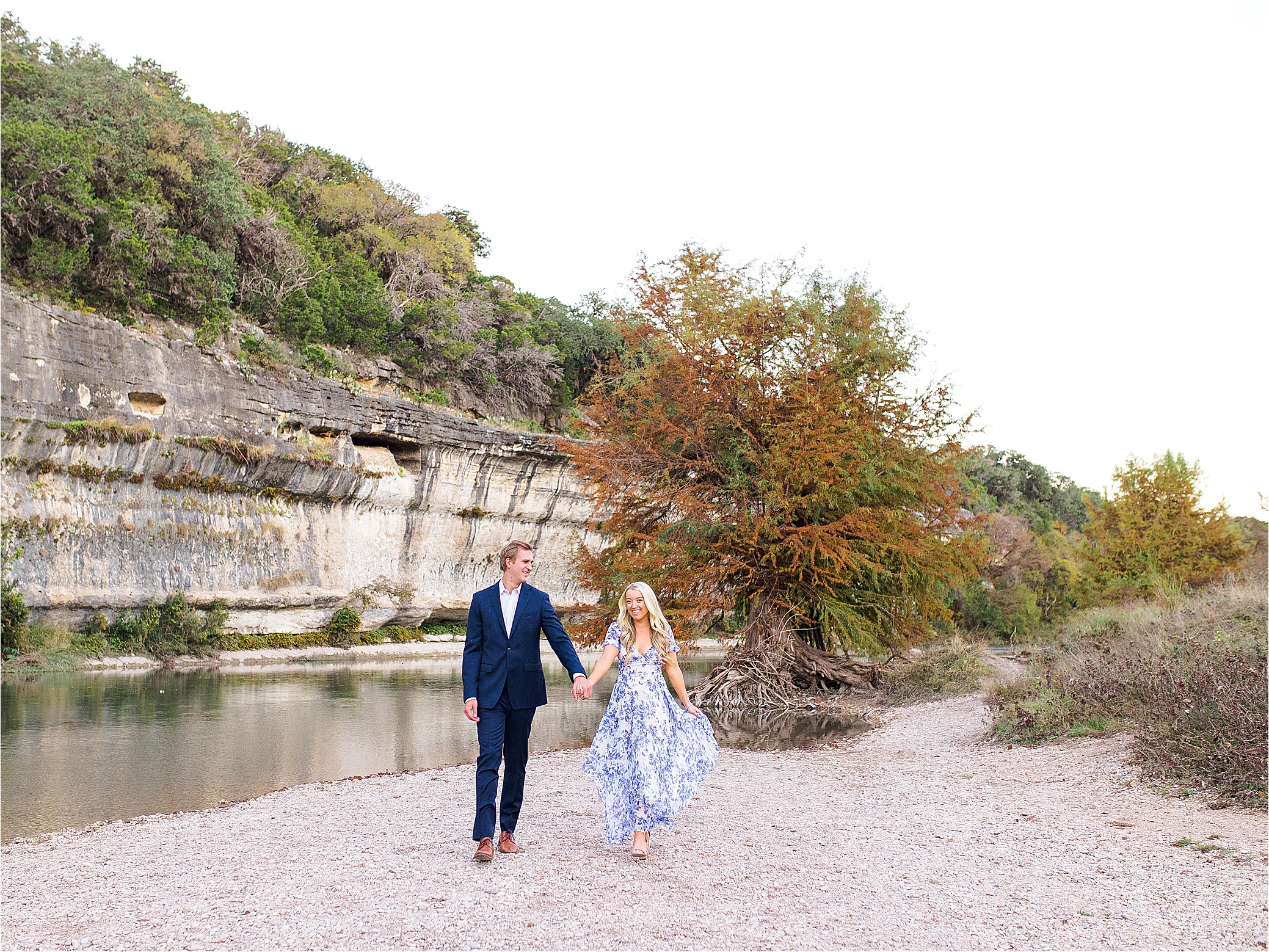 An engaged couple holds hands and walks toward the camera with her blue and white dress swaying in front of a river and cliffs at Guadalupe River State Park near Boerne, Texas for their Hill Country Engagement Session 