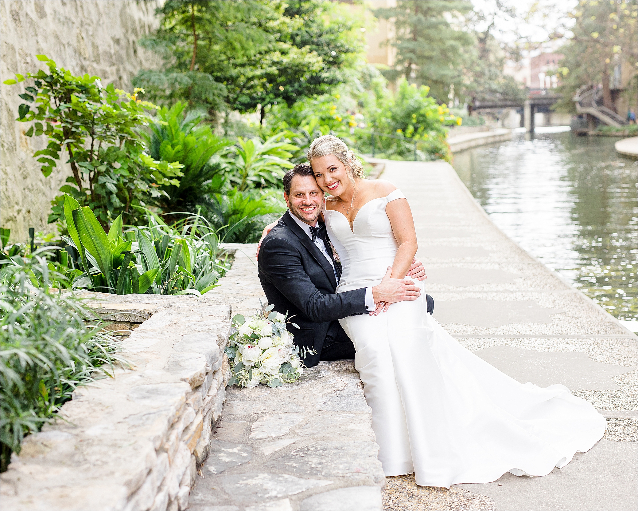A bride sits on her grooms lap and hugs him in front of the San Antonio Riverwalk for their wedding day portraits with Jillian Hogan Photography 