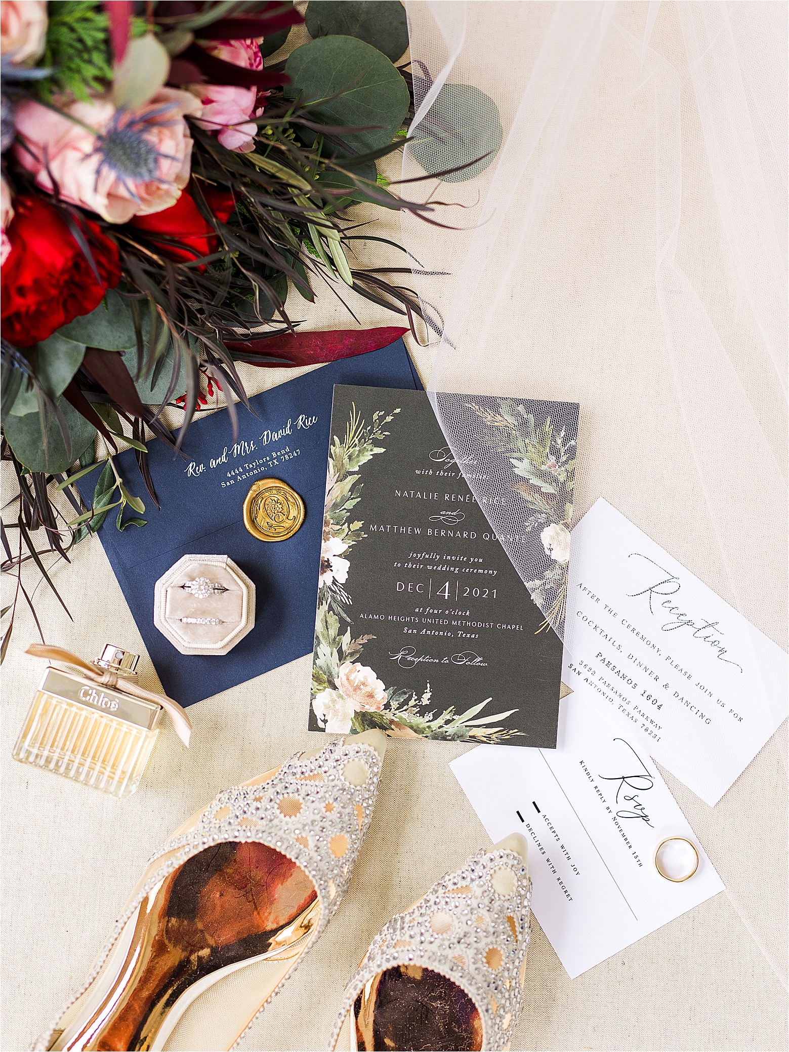 An invitation suite with red and pink flowers, golden wedding shoes, chloe perfume and a navy flat lay and ring box for a San Antonio Winter Wedding