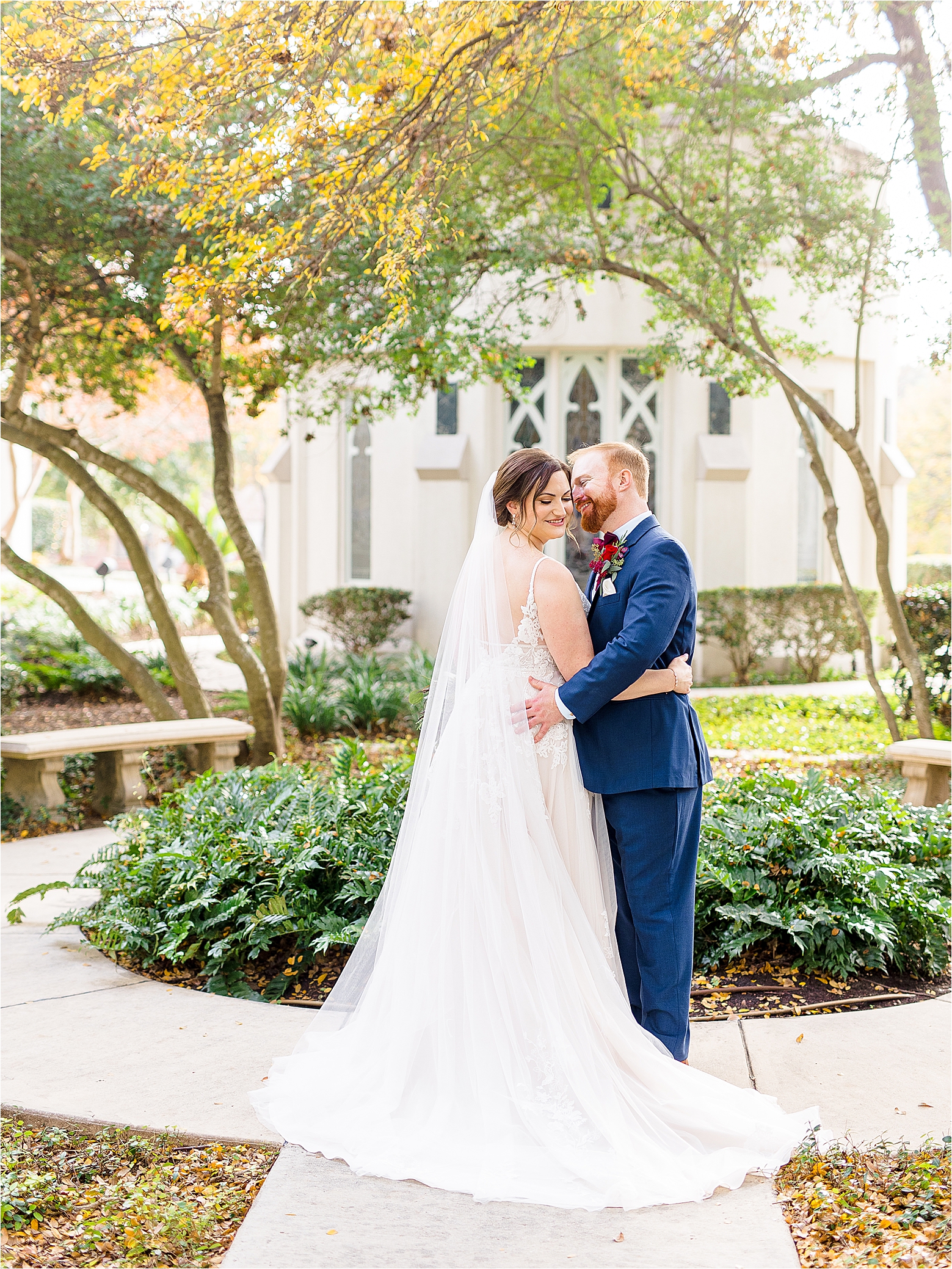 A groom places his hand on his brides waste and whispers to her as she looks down over her shoulder in front of greenery at Alamo Heights United Methodist Church in San Antonio, Texas 
