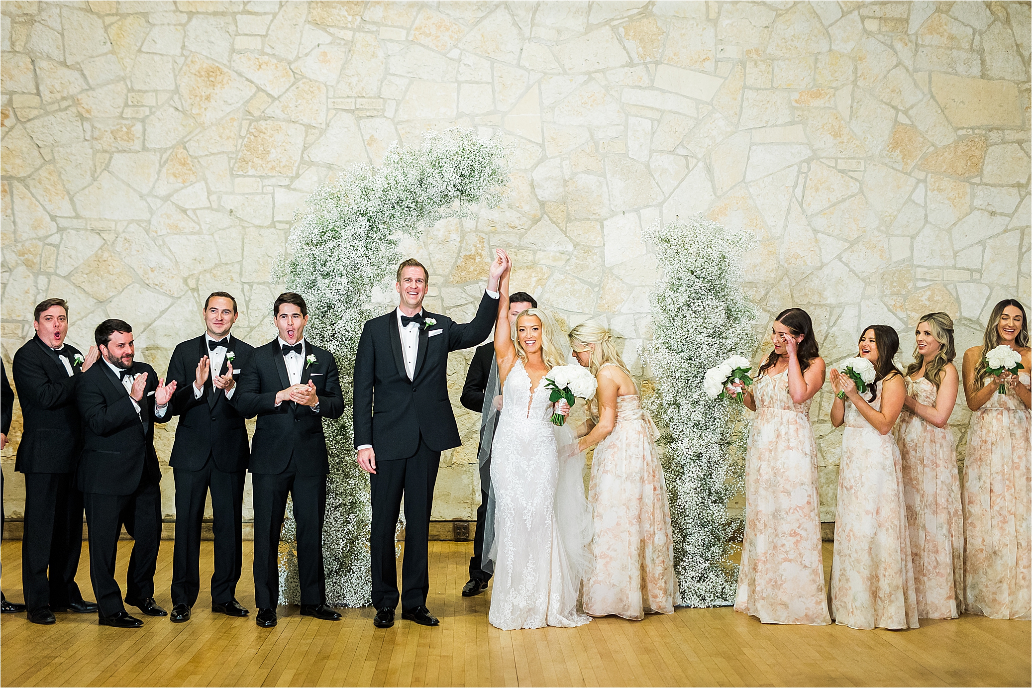 A bride and groom celebrate and hold their hands up as they are pronounced husband and wife in front of an arch of baby's breath and a stone wall at Brodie Homestead in Austin, Texas. 