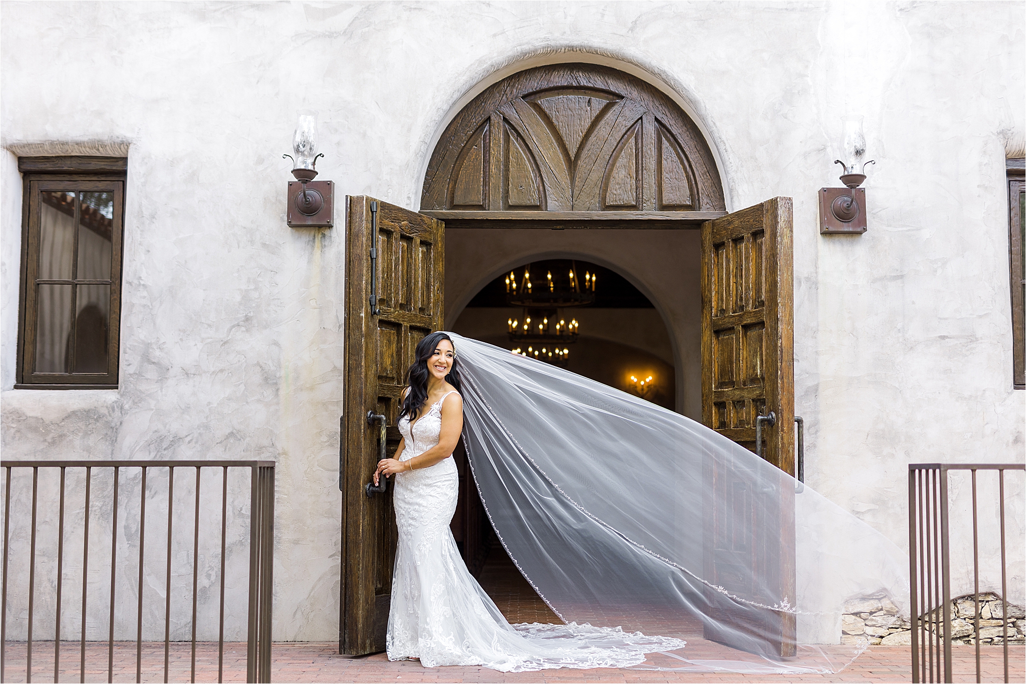 A bride smiles back over her shoulder as her veil blows in the wind in front of an old brown door at a Mission Venue in San Antonio Texas during her bridal session with Jillian Hogan