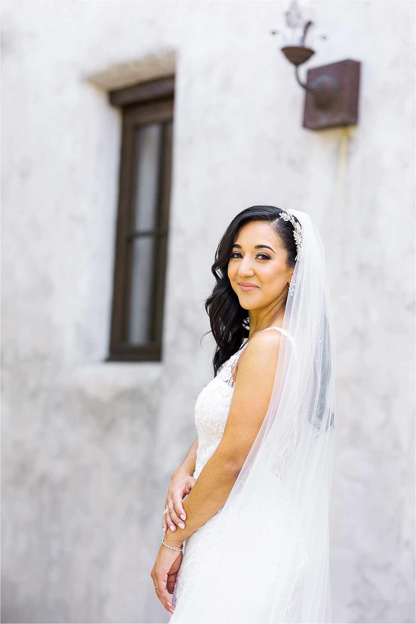 A bride smiles back at the camera with her veil over her shoulders and in front of a white mission building with San Antonio Wedding Photographer Jillian Hogan Photography