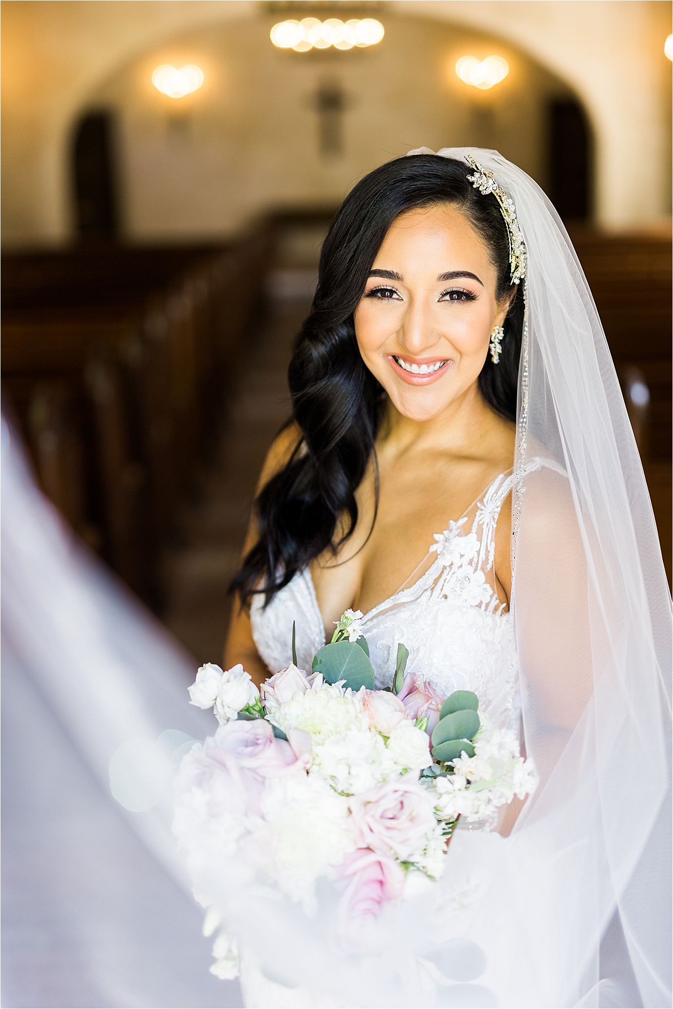 A bride smiles with inside a mission chapel as her veil swoops in front of her holding a bouquet of pink and white flowers during her bridal session at Lost Mission in Bulverde, Texas
