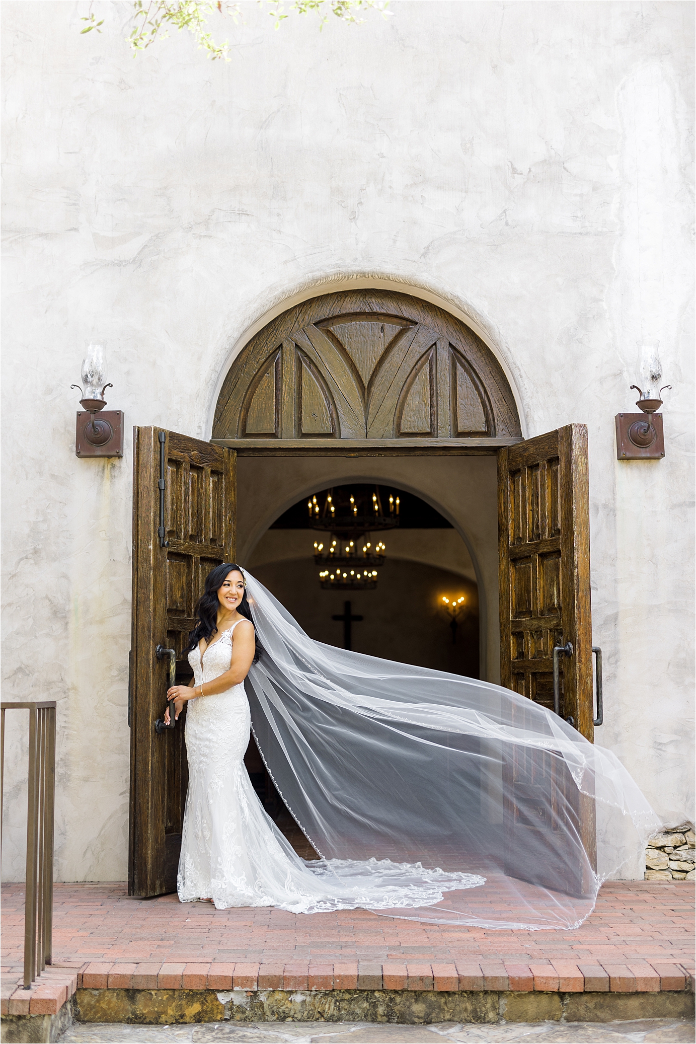 A bride looks off to the side as her veil blows in the wind in front of big, brown doors at her Mission Venue in the Texas Hill Country during her bridal Portraits