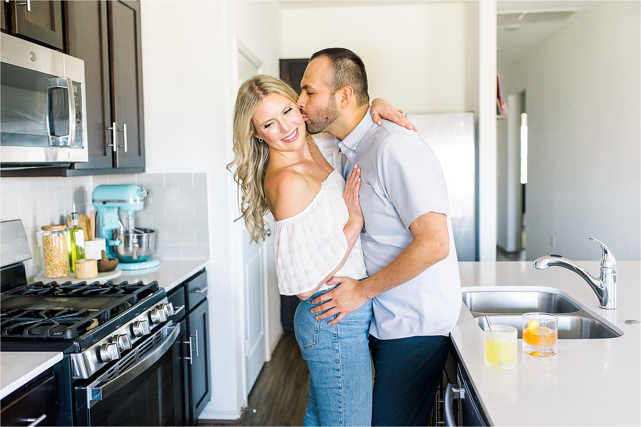 A guy kisses his fiance on the cheek in their kitchen for their at home engagement session in Austin, Texas