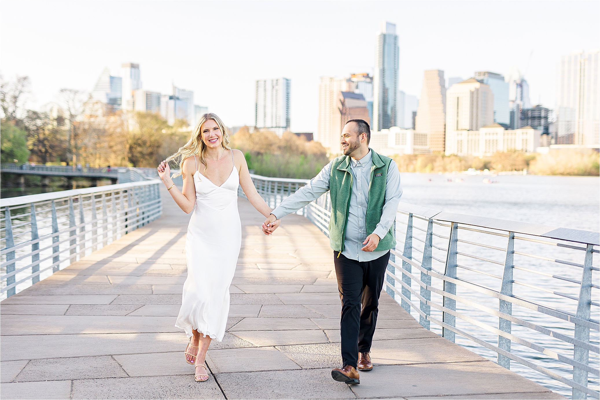 A couple playfully holds hands walking down boardwalk in Austin with skyline behind them for their engagement session with Jillian Hogan Photography