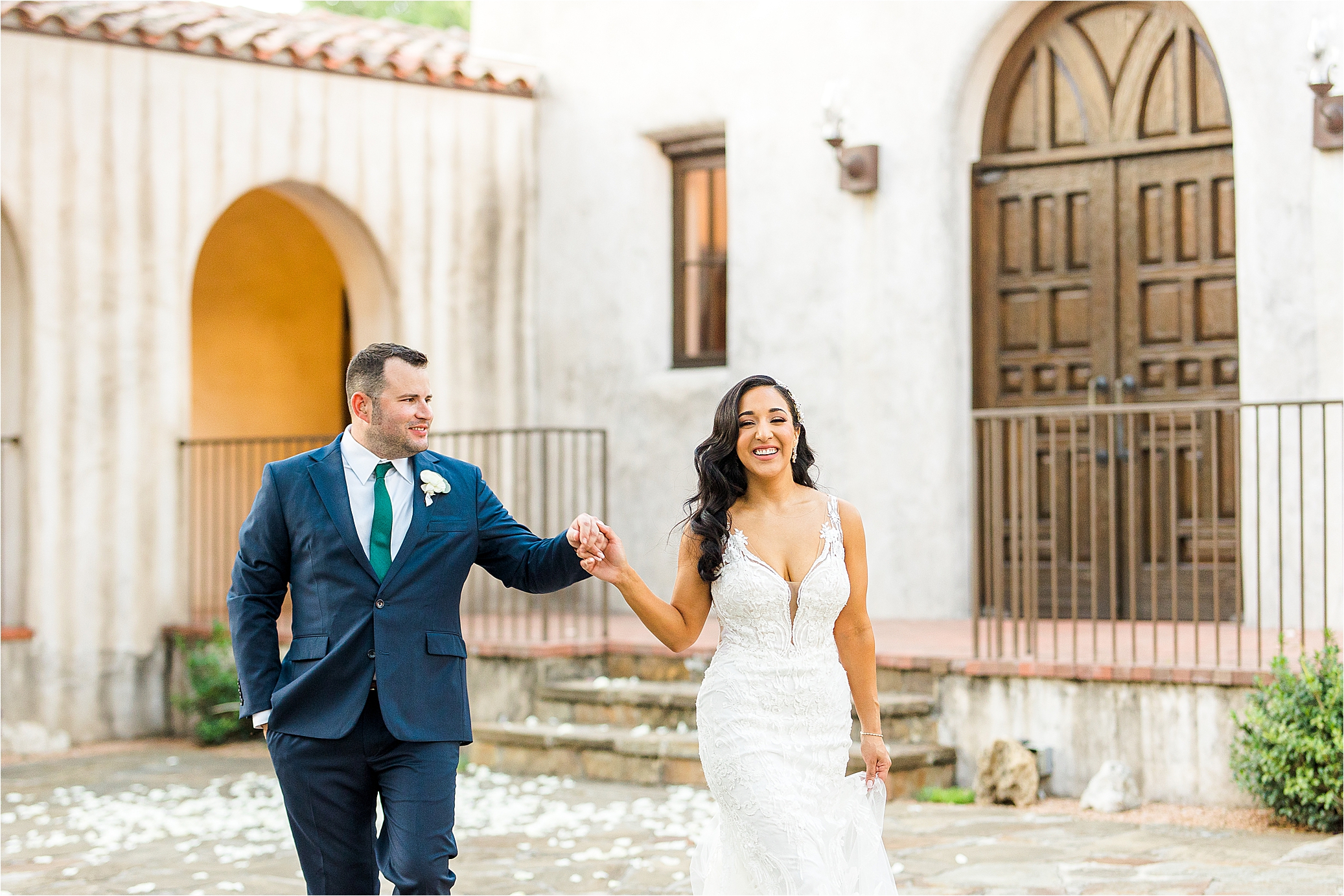 A newlywed couple walks toward the camera and smiles in front of Mission Style Chapel near San Antonio, Texas 
